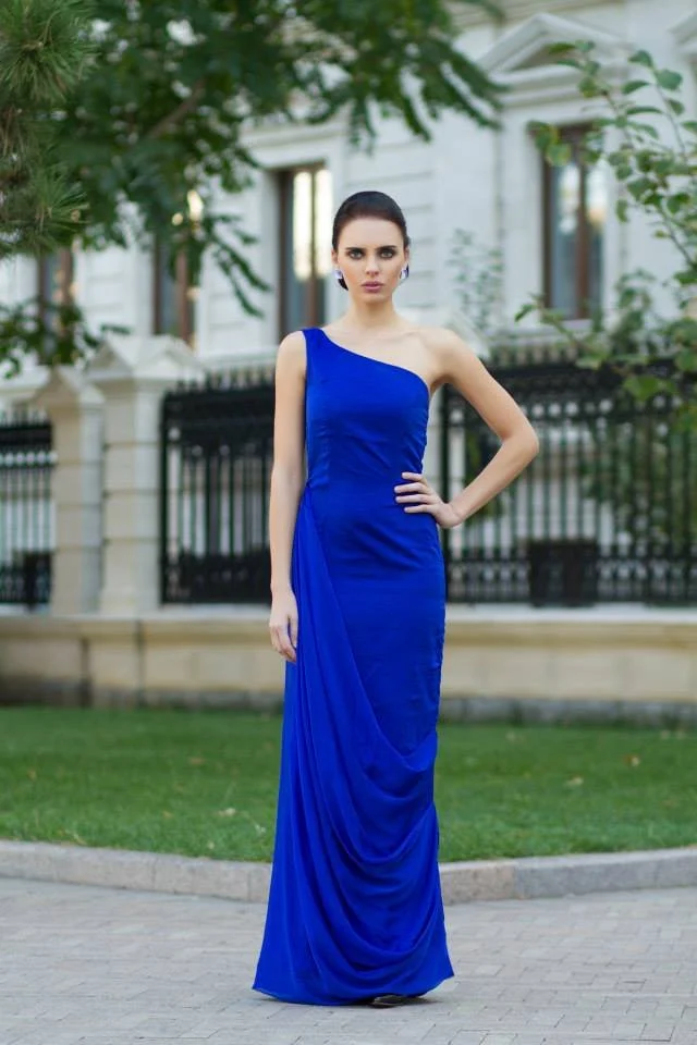 A woman in a royal blue evening gown standing outdoors on a paved path with a white building and black fence in the background.