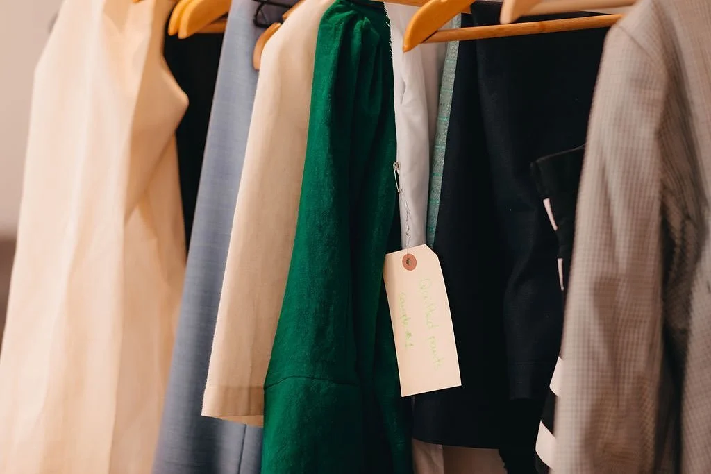 Clothing rack with various dresses and blouses hanging on wooden hangers, including beige, blue, green, black, and checked patterns, with a tag attached to one of the garments.