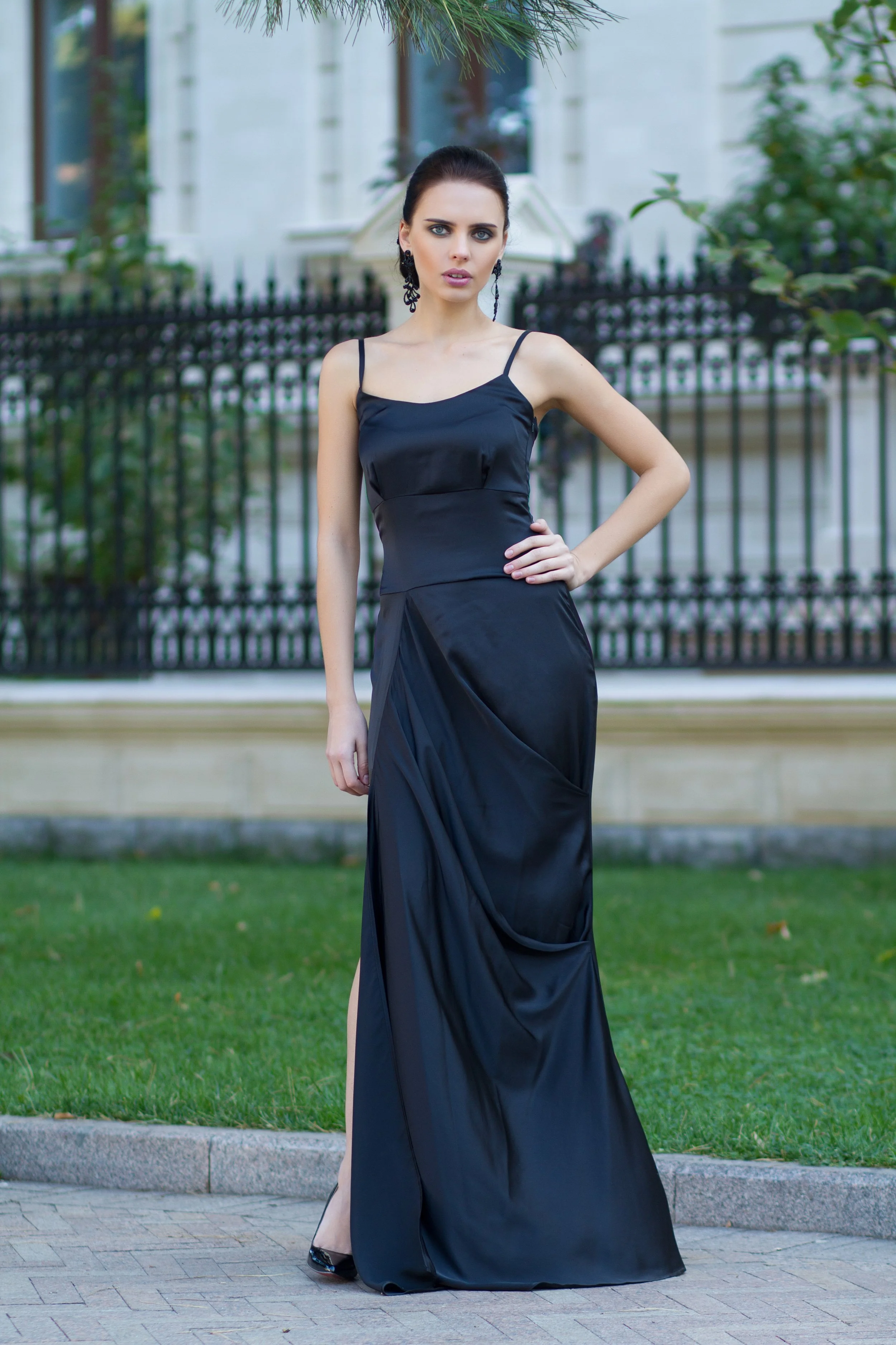 A woman in a long black satin gown standing outdoors on a sidewalk with a green lawn and a white building in the background.