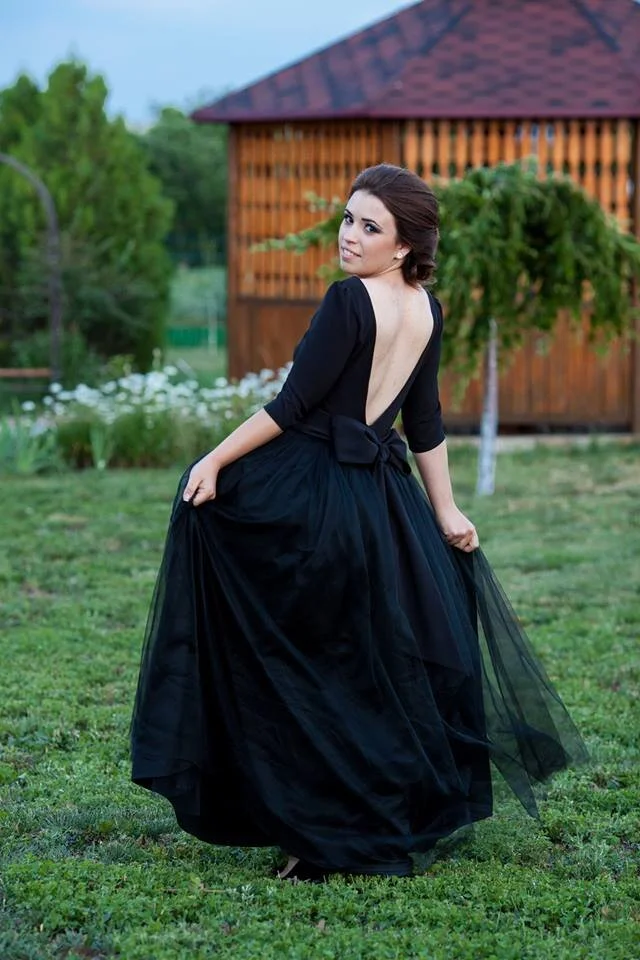 A woman in a black dress with a low back, holding the skirt, outdoors on a grassy area with trees and a wooden building in the background.