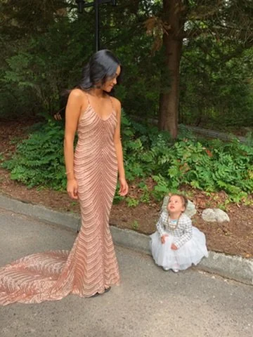 A woman in a long, gold, sequin dress looking down at a young girl in a white dress and gray cardigan sitting on the sidewalk, with greenery in the background.