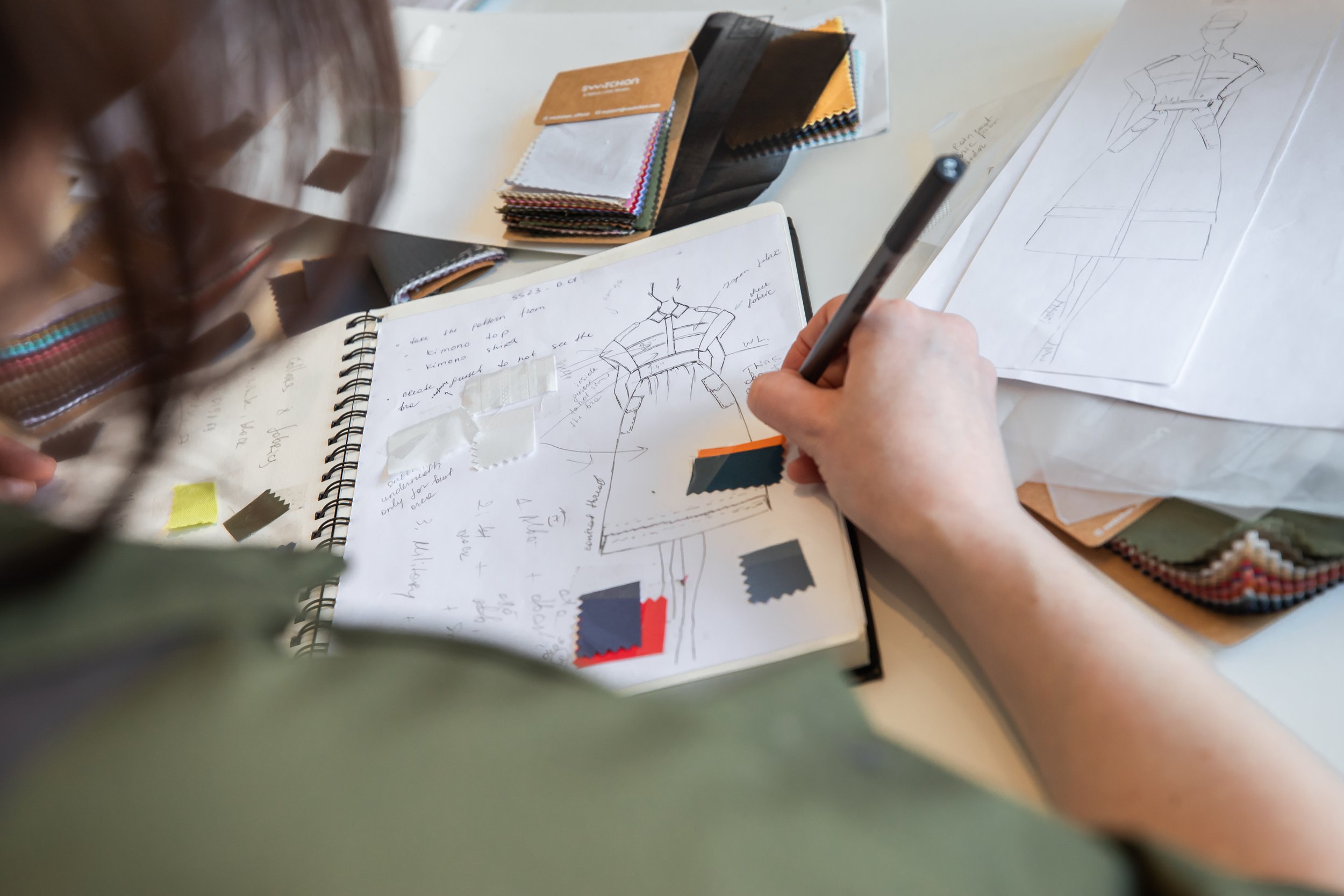 Person designing a dress sketch with fabric swatches, color samples, and pattern papers on a desk.