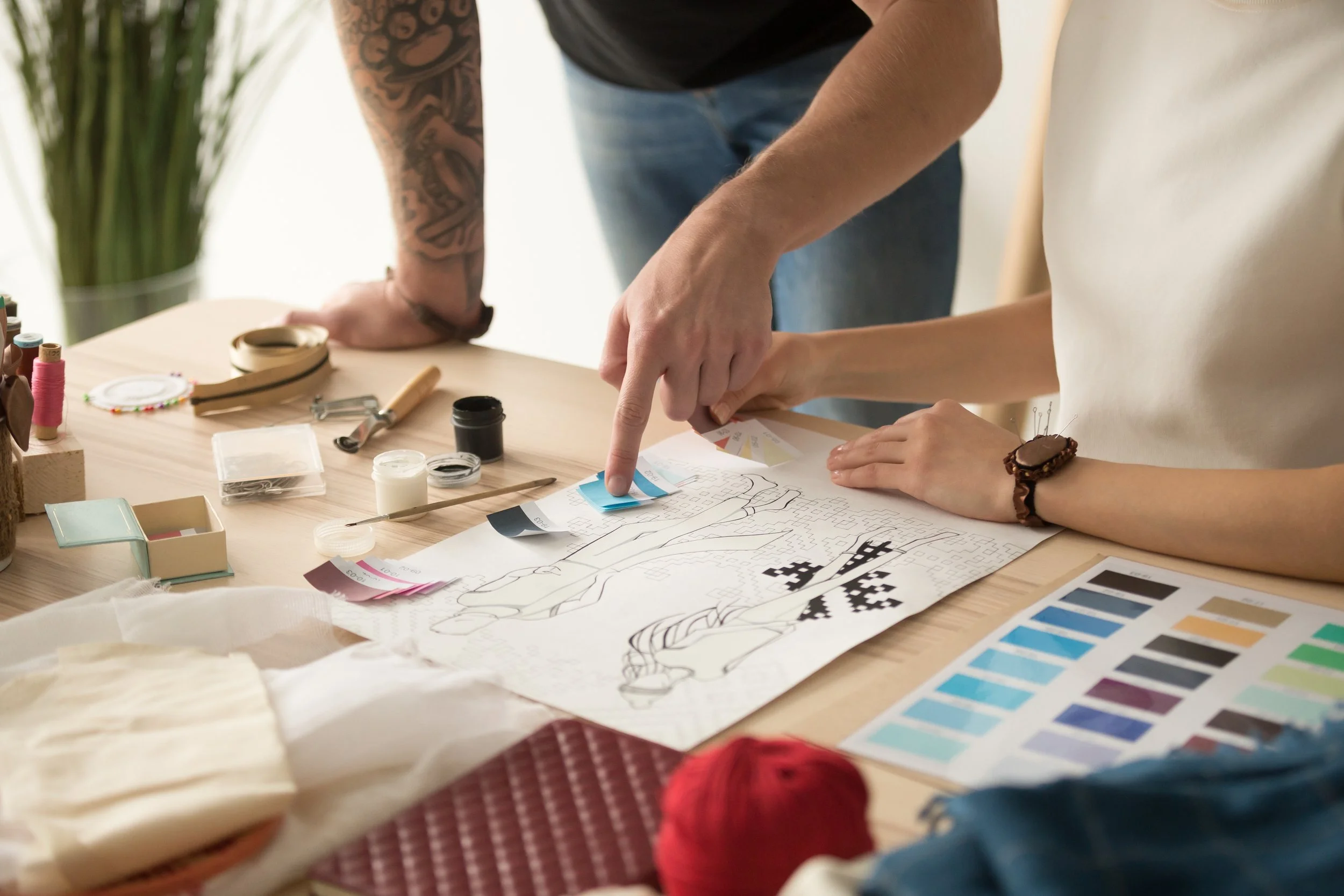 Two people engaged in a craft project at a table, with various tools, colors, and fabric swatches, working on a design or sewing pattern.