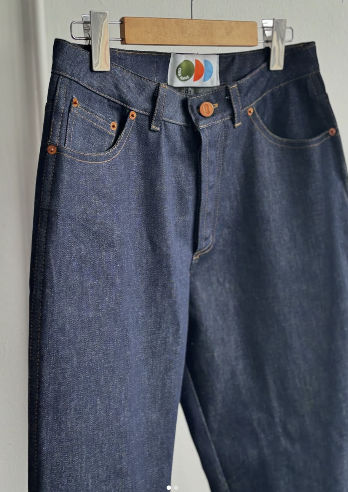Pair of dark blue denim jeans hanging on a wooden hanger against a light-colored wall.