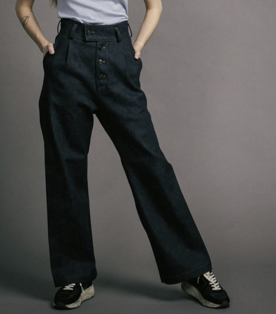 Person wearing high-waisted black pants, gray shirt, and black sneakers, posing with hands in pockets against a gray background.