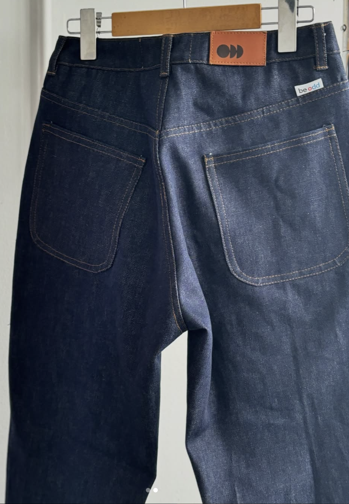 Back view of a pair of dark blue jeans hanging on a wooden hanger with metal clips.