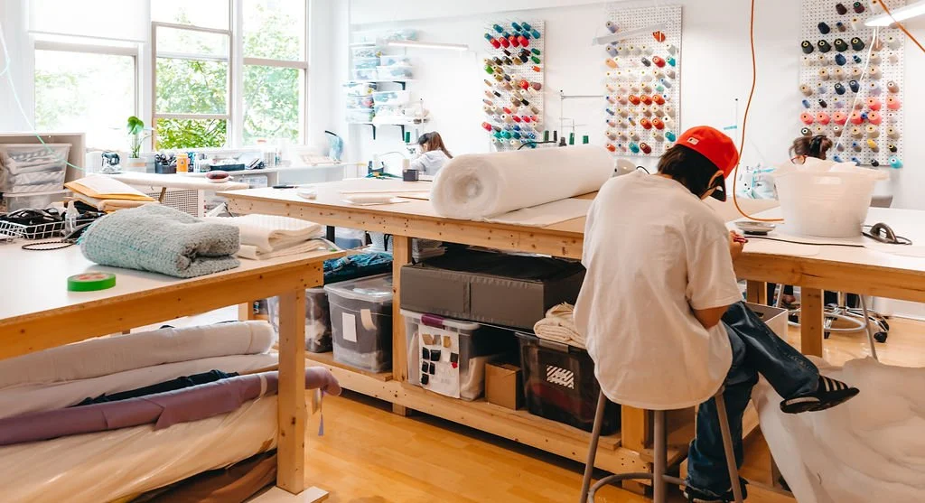 Art workspace with sewing materials and fabric on wooden tables, people working at sewing machines, and colorful spools of thread on the wall.
