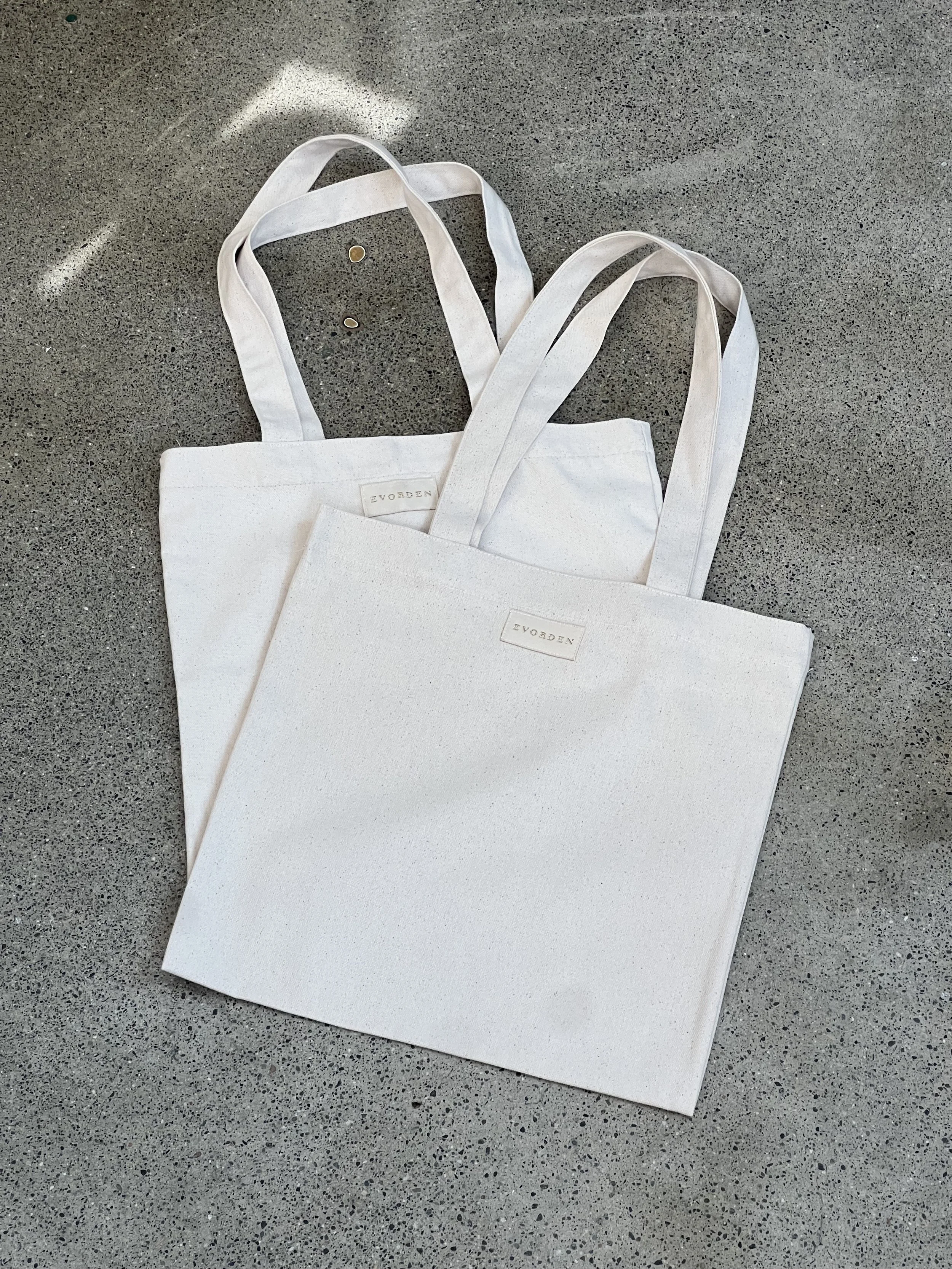 Two white canvas tote bags laying on a gray speckled surface, with small gold coins beside them.