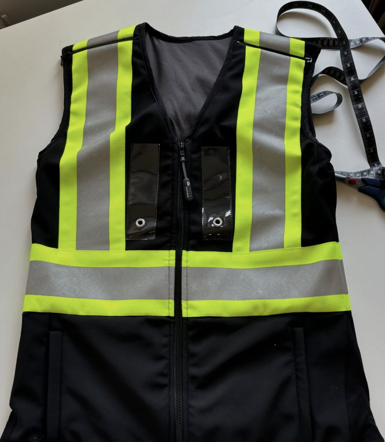 High-visibility reflective safety vest with zipper, black and gray fabric, and attached clear plastic pouches for identification or tools, laid on a white surface next to a measuring tape and scissors.