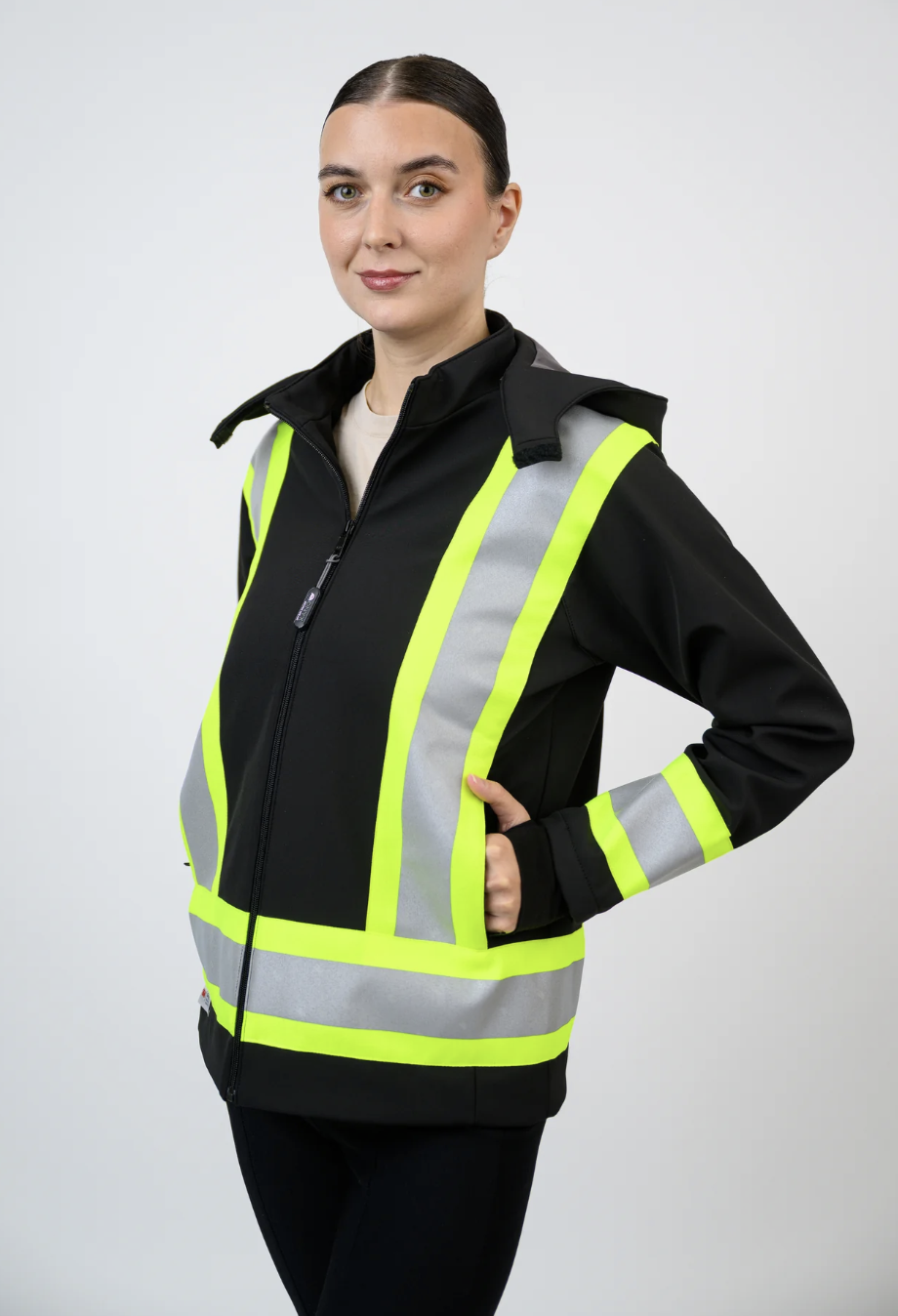 A woman wearing a black jacket with reflective yellow and gray safety stripes, standing against a plain light gray background.