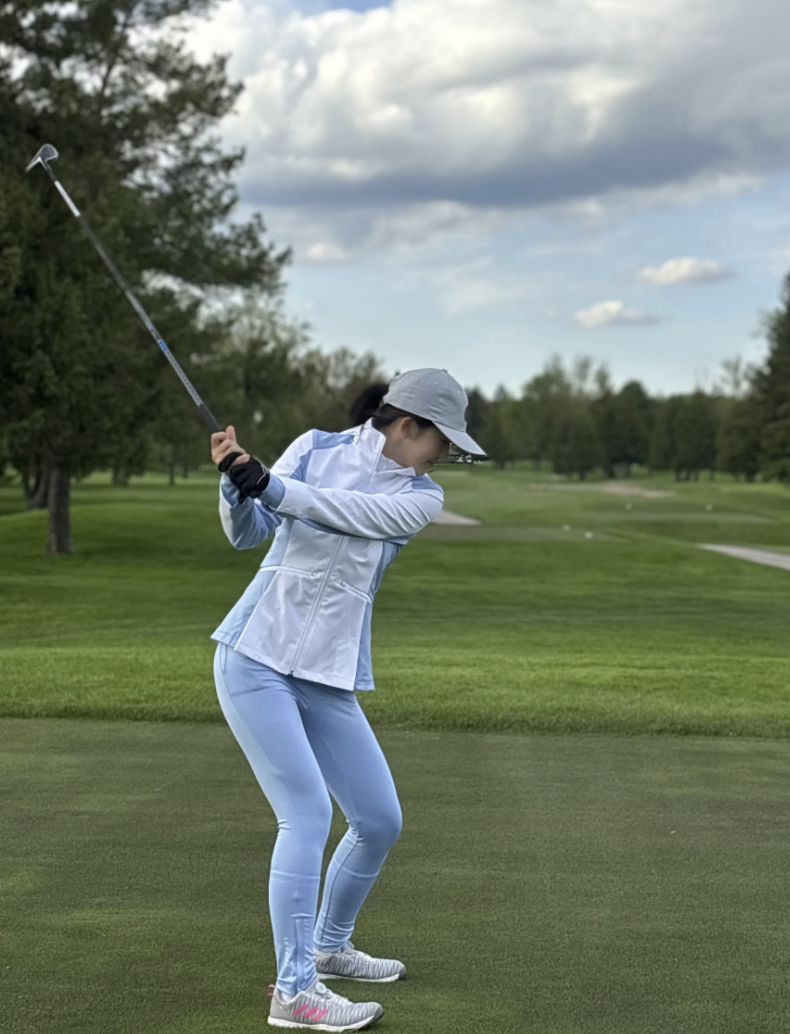 A woman in white sportswear and a gray hat is swinging a golf club on a golf course with green grass, trees, and a partly cloudy sky.