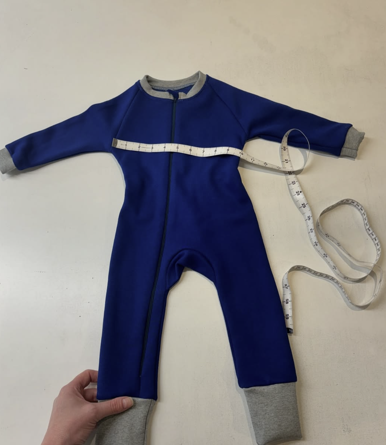 Blue children's jumpsuit with gray cuffs and zipper, measuring tape laid across it, on a white surface.