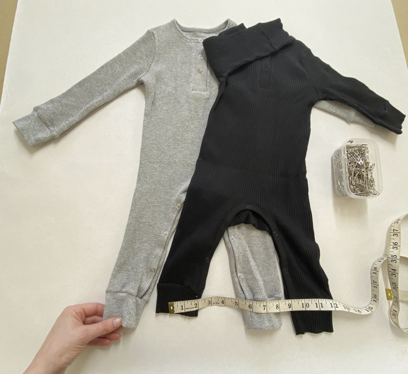 Two baby onesies, one grey and one black, laid out on a white surface with a measuring tape and a container of safety pins nearby.