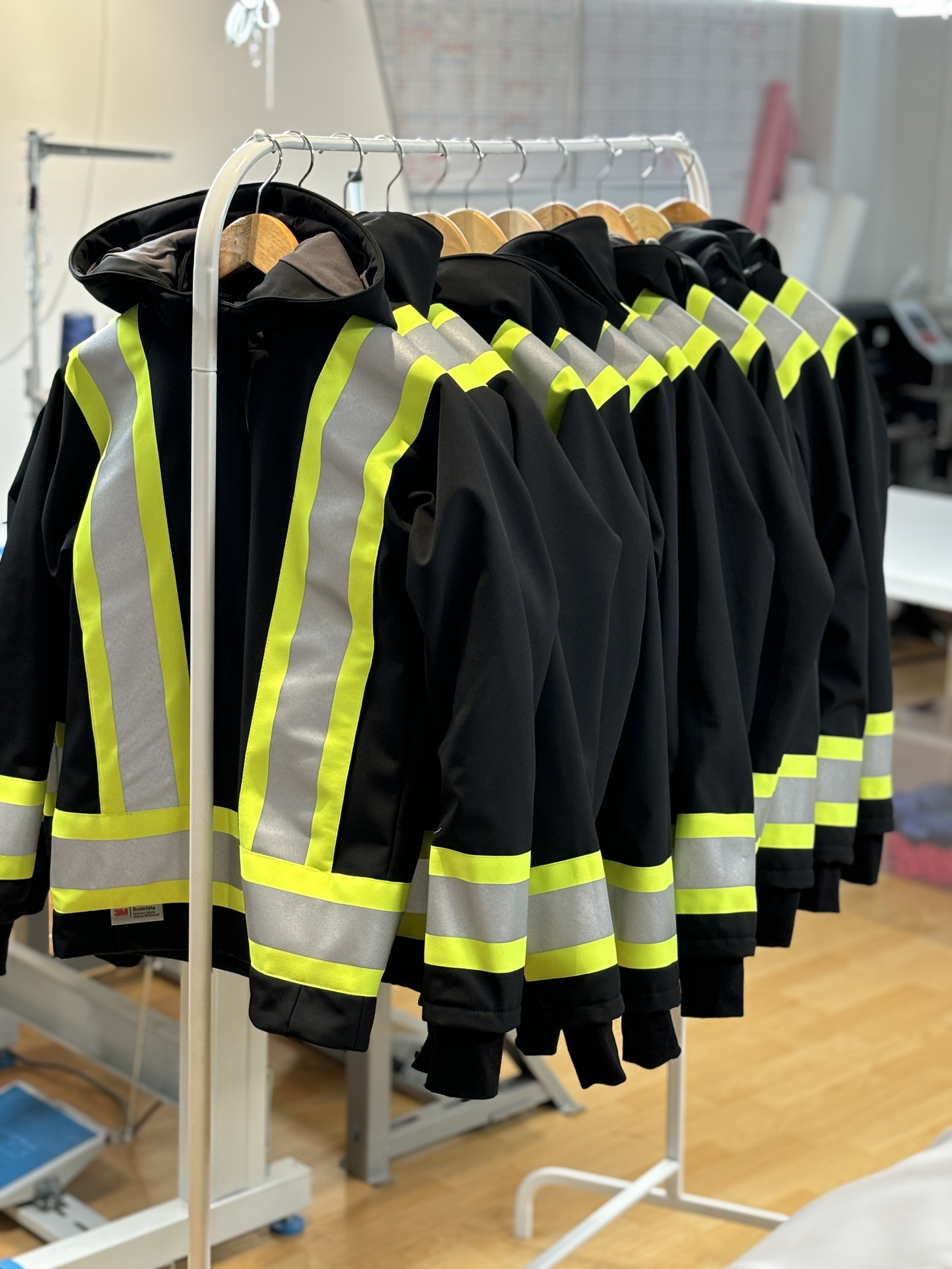 Black firefighter jackets with yellow reflective stripes hanging on a white clothing rack.
