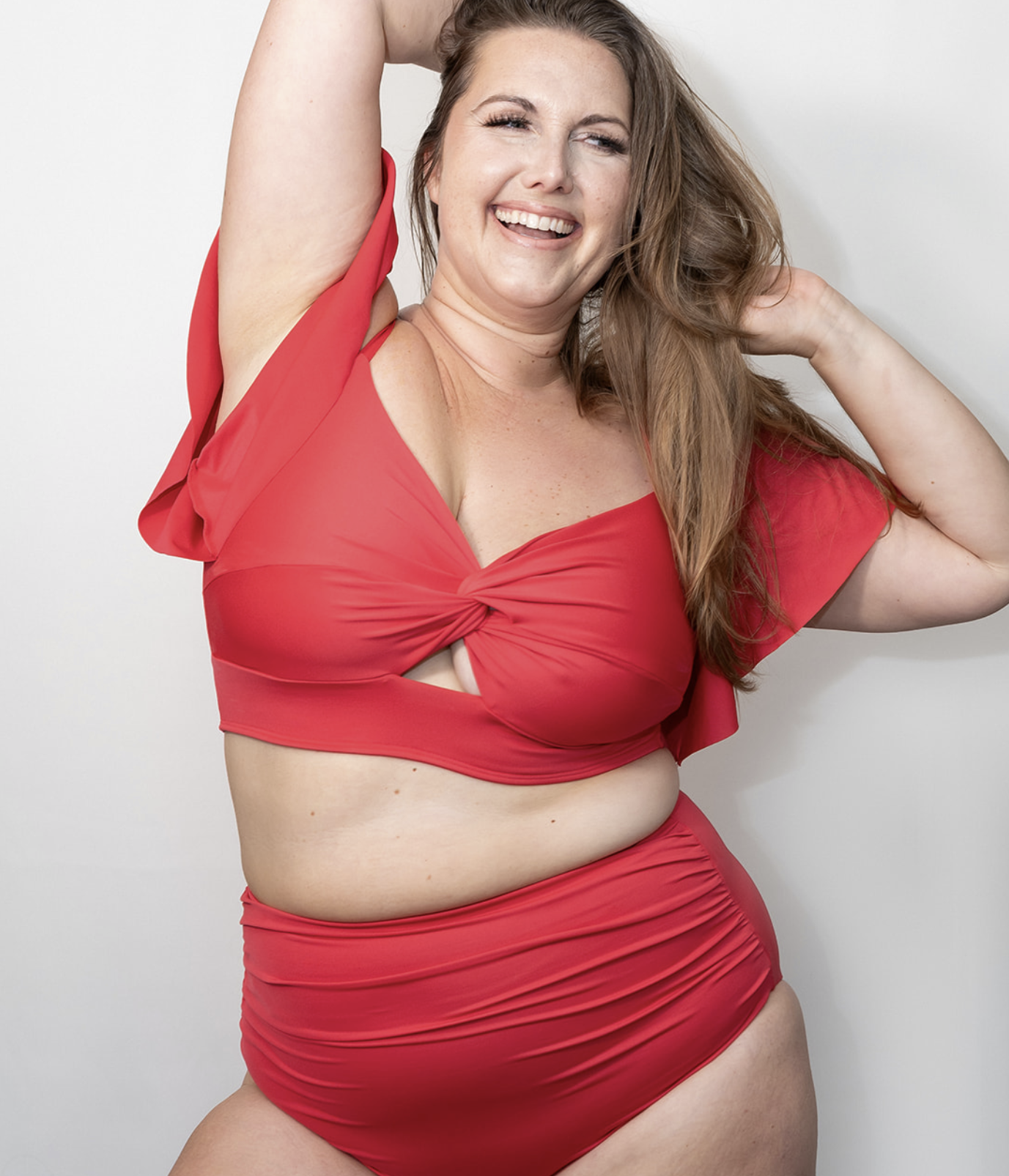 A joyful woman posing in a red bikini with cutouts, smiling and holding her hair back, standing against a plain white background.