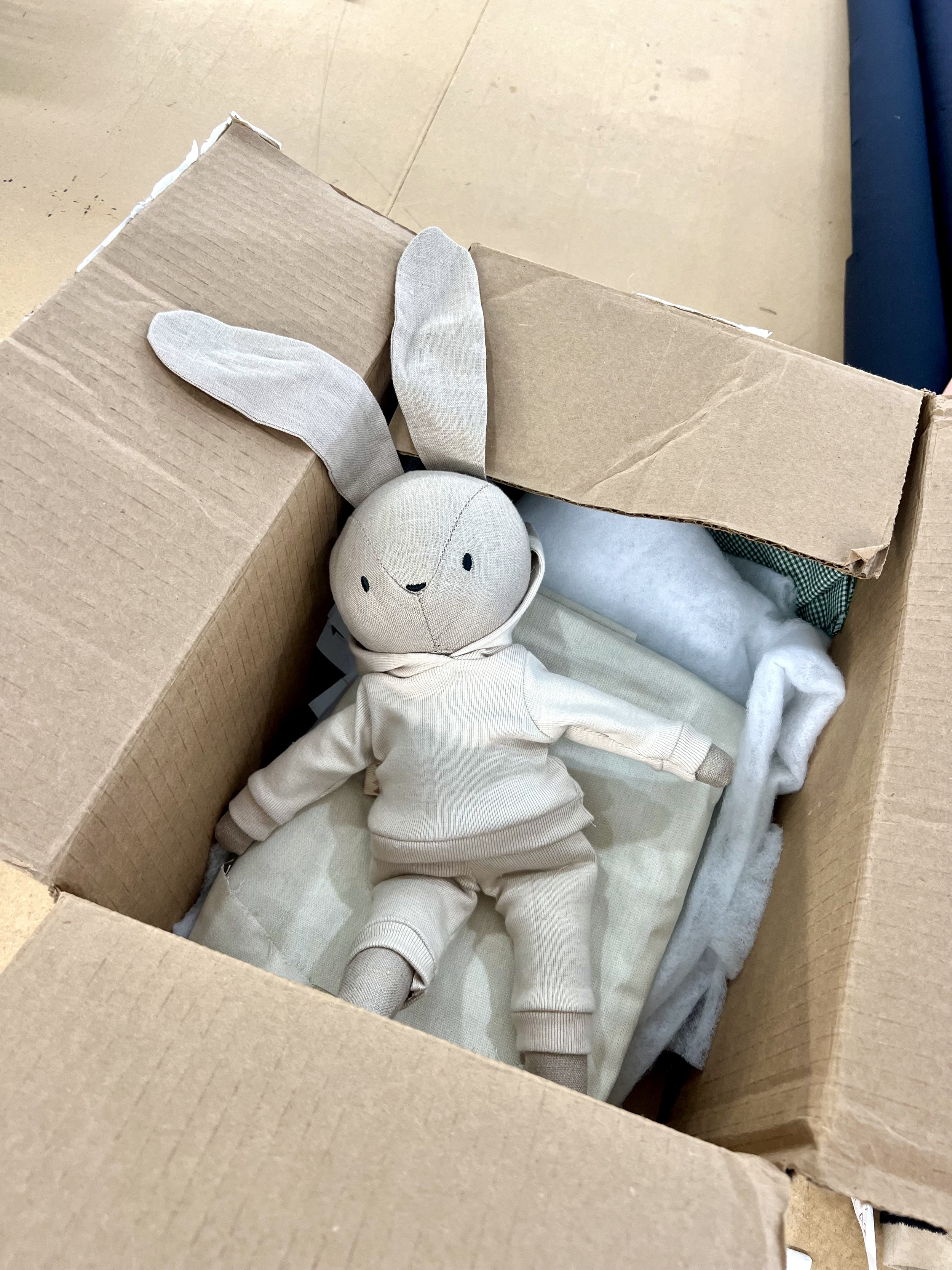 A stuffed rabbit toy with long ears, wearing a cream-colored hoodie and pants, lying inside a cardboard box along with some white and light-colored fabric or blankets.
