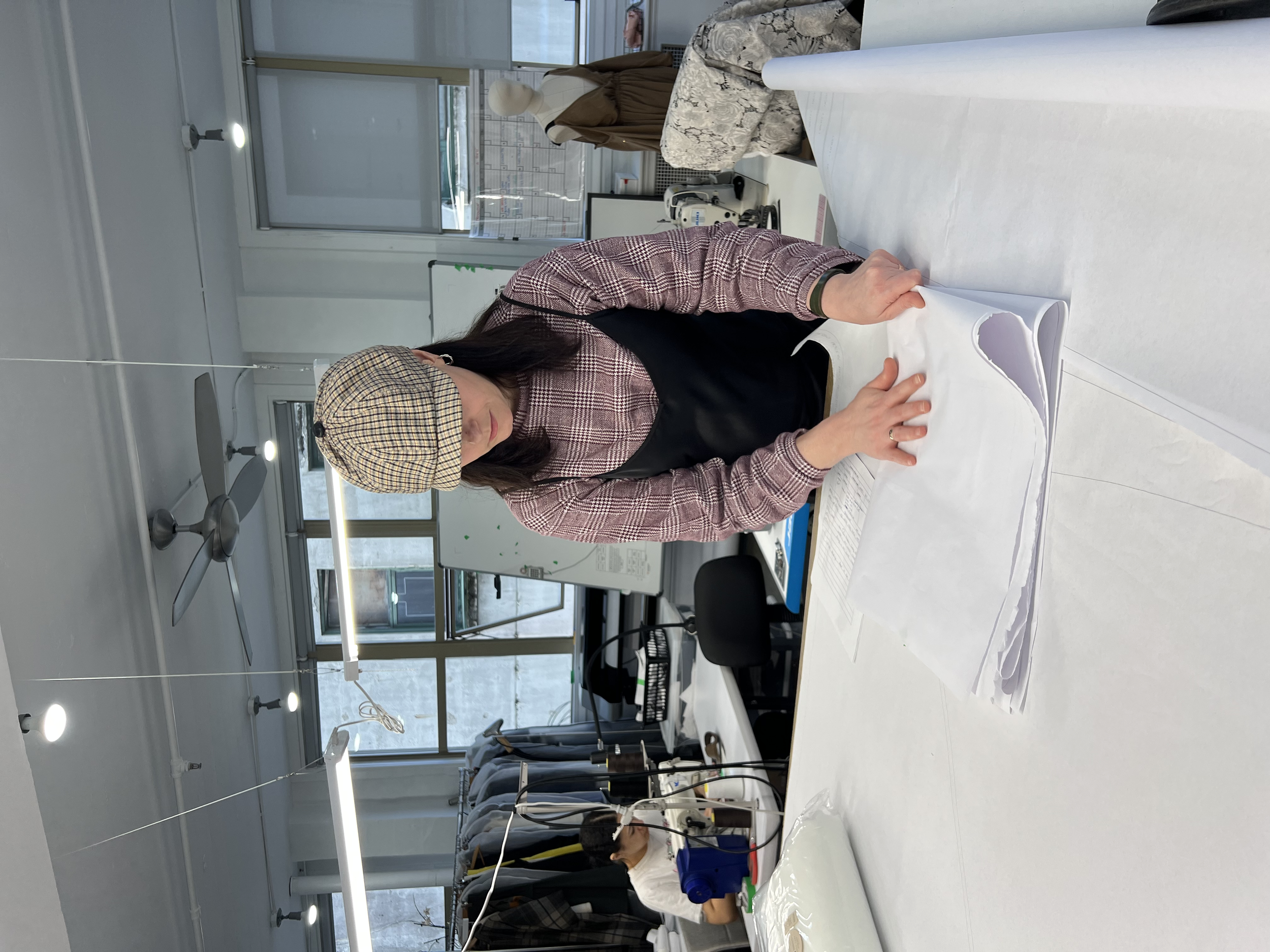 Woman in a plaid cap and apron working with fabric in a sewing or textile workshop.