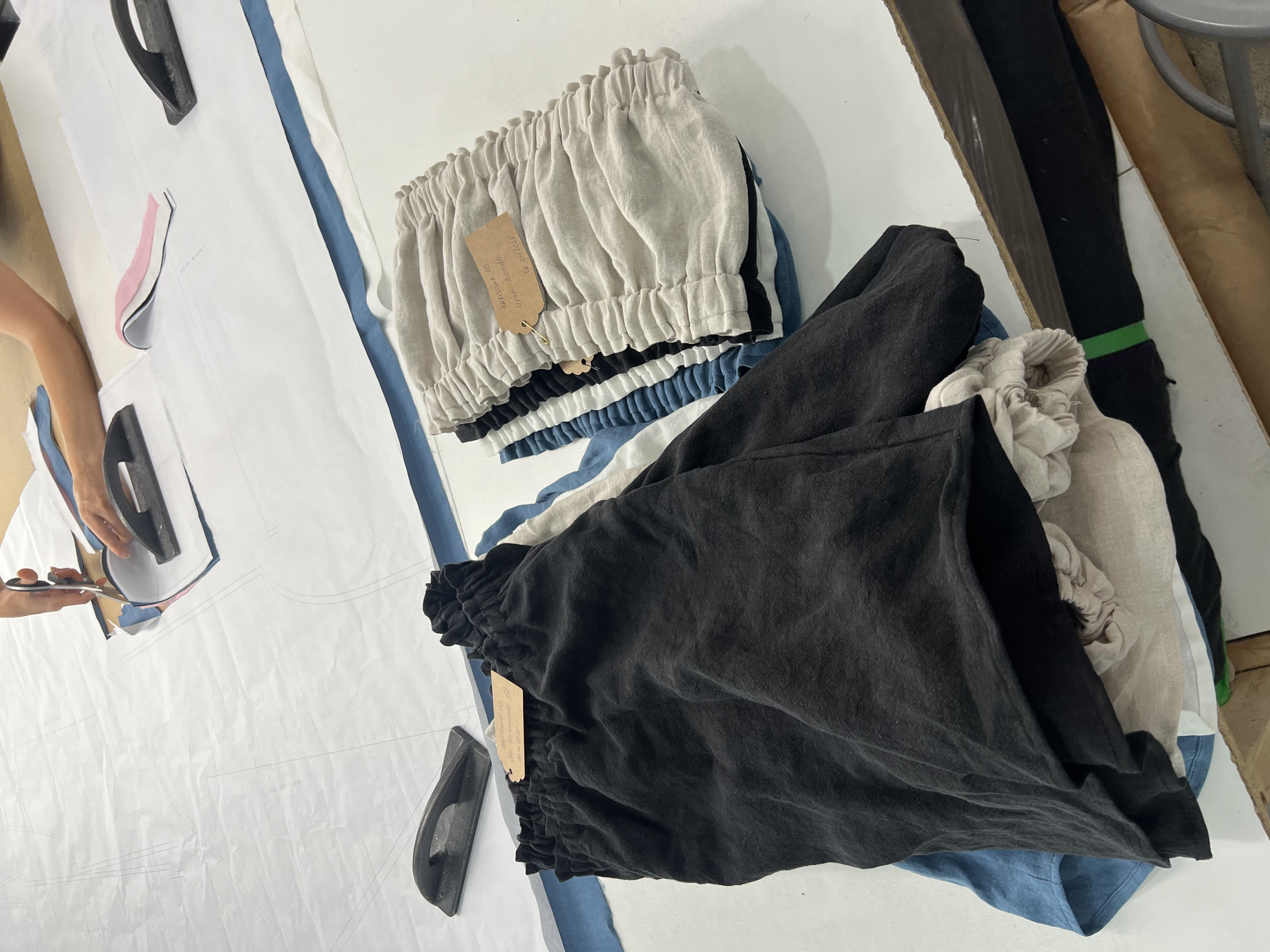 Clothes in various colors, including black, beige, and blue, laid out on a table with tags attached, while a person in the background uses a rotary cutter on fabric.