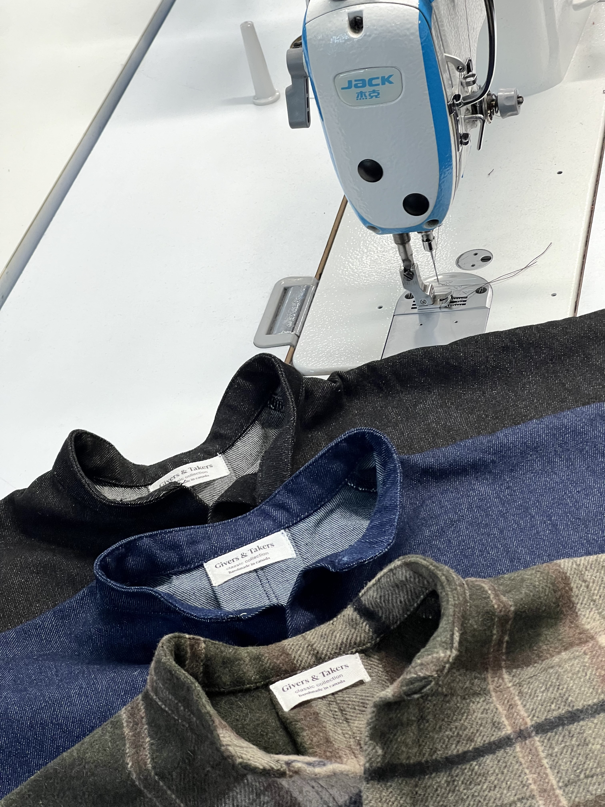 A sewing machine stitching fabric shirts in black, blue, and plaid, with focus on the shirts' collars and labels, on a white sewing table.