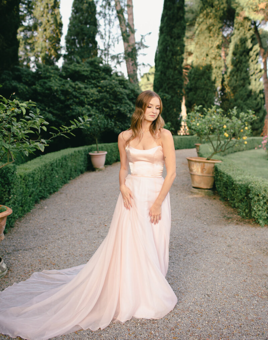 A woman in an elegant light pink gown with a train standing in a garden with lush green trees and potted plants.