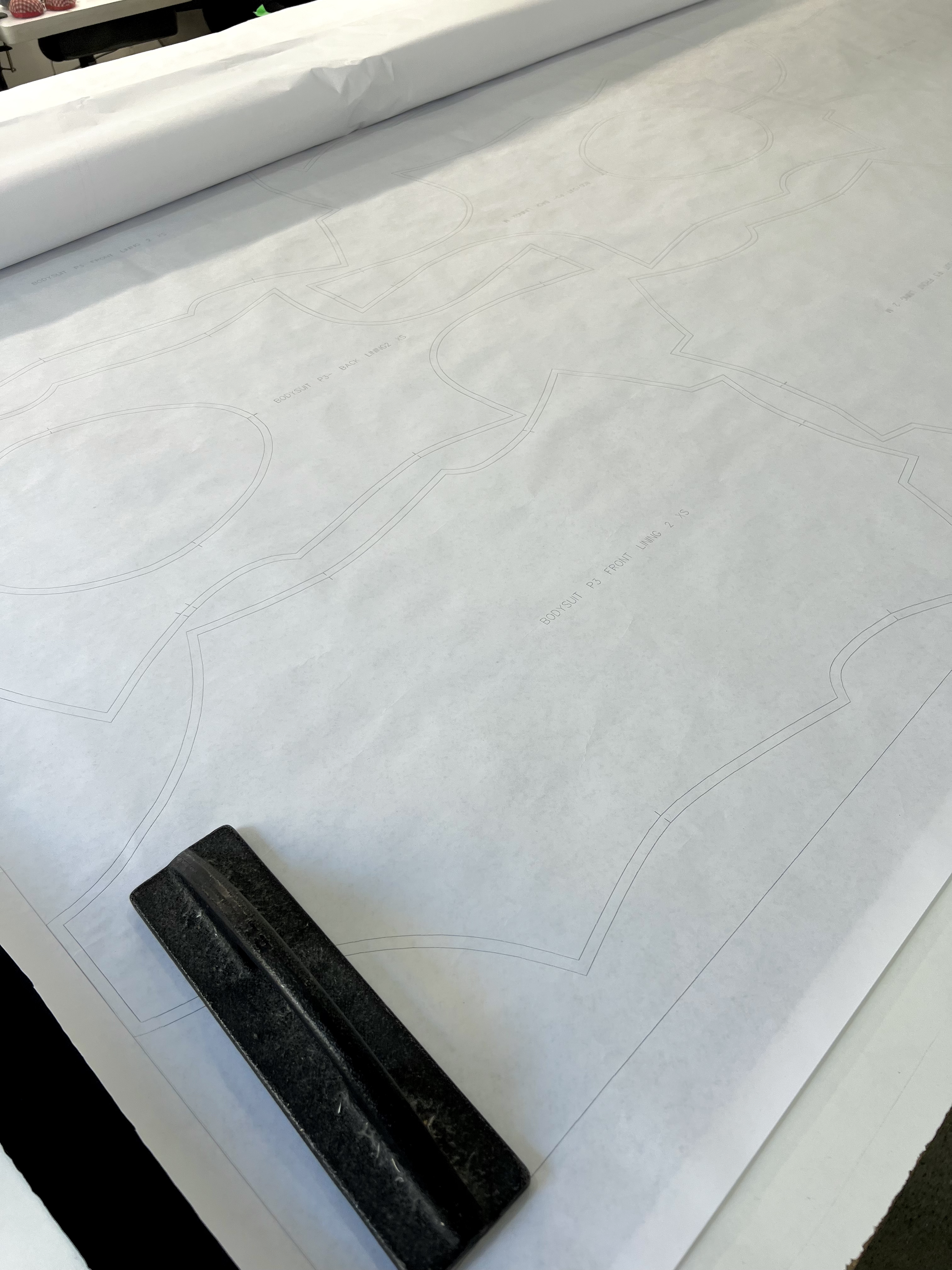 Large sheet of pattern paper with outline designs, a black weight placed on the bottom left corner, and some faint printed text indicating pattern parts.
