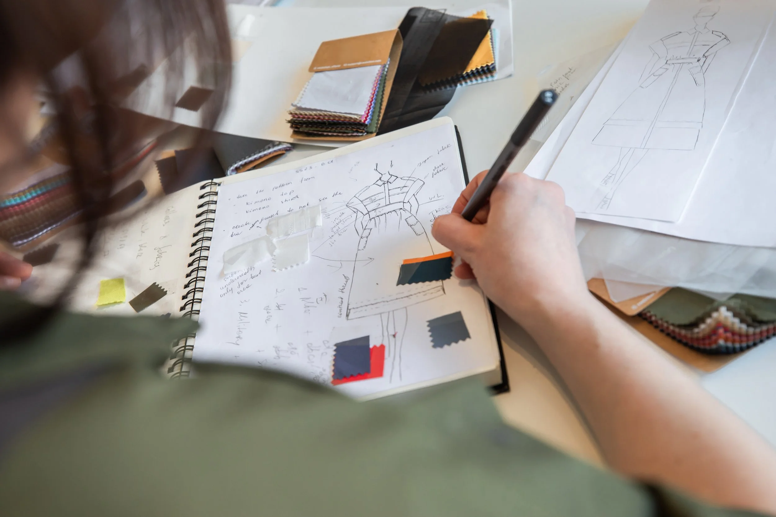 A person is sketching a fashion design in a notebook, surrounded by fabric swatches, sketches, and color samples on a workspace table.
