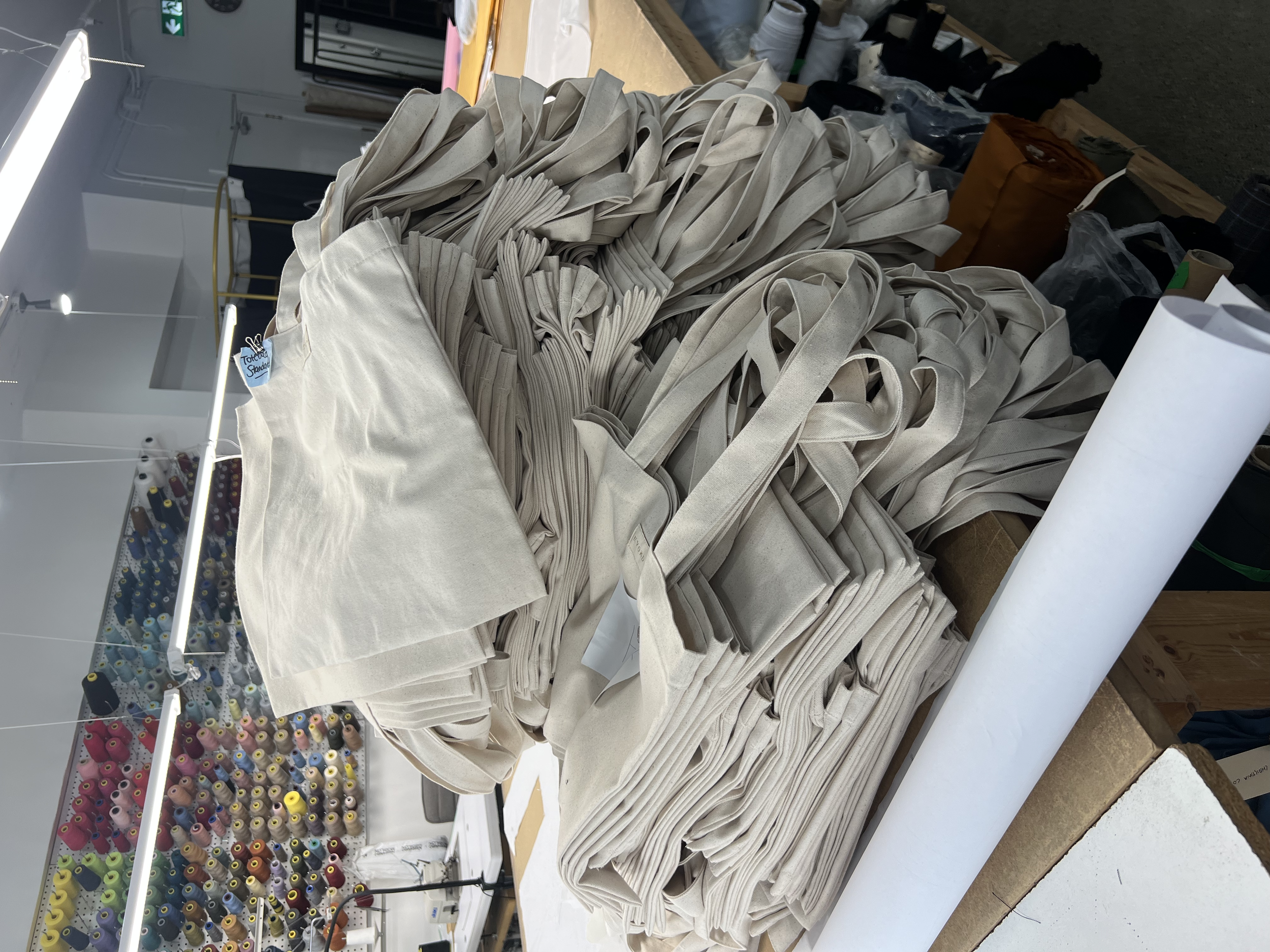 Stack of natural-colored canvas tote bags and fabric rolls on a worktable in a sewing or craft workshop.