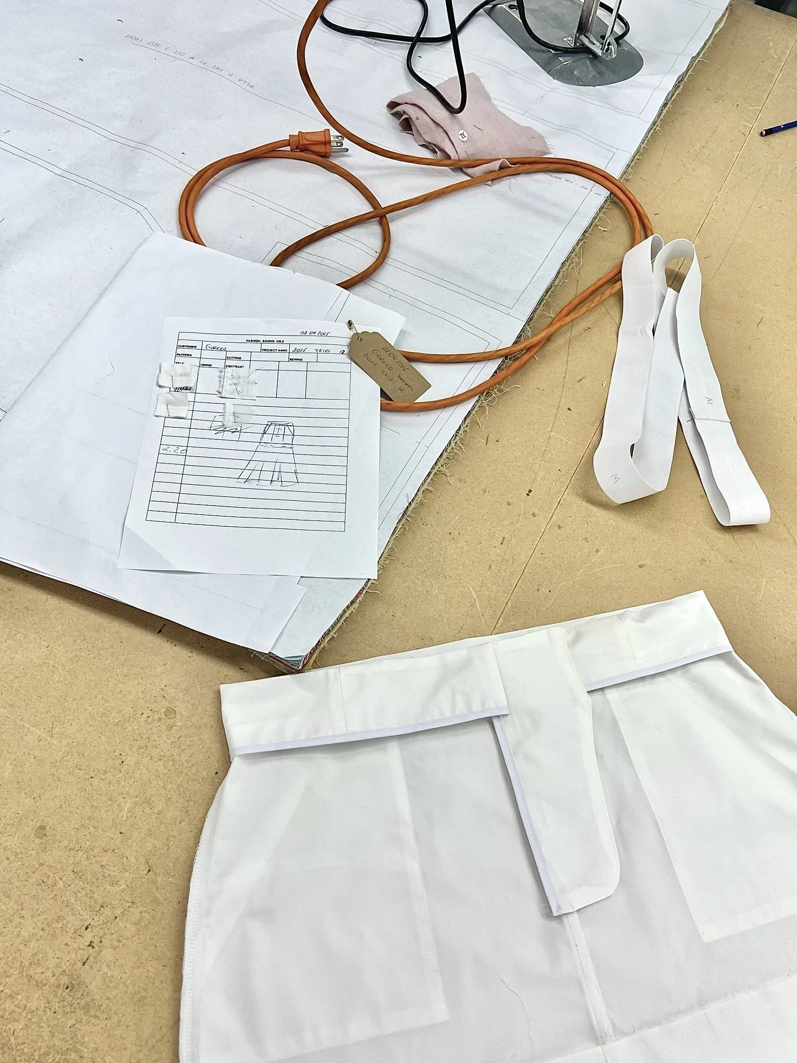 Worktable with electrical cords, design plans, a fabric piece with pockets, a white apron, and a pattern sketch, indicating a sewing or tailoring workspace.