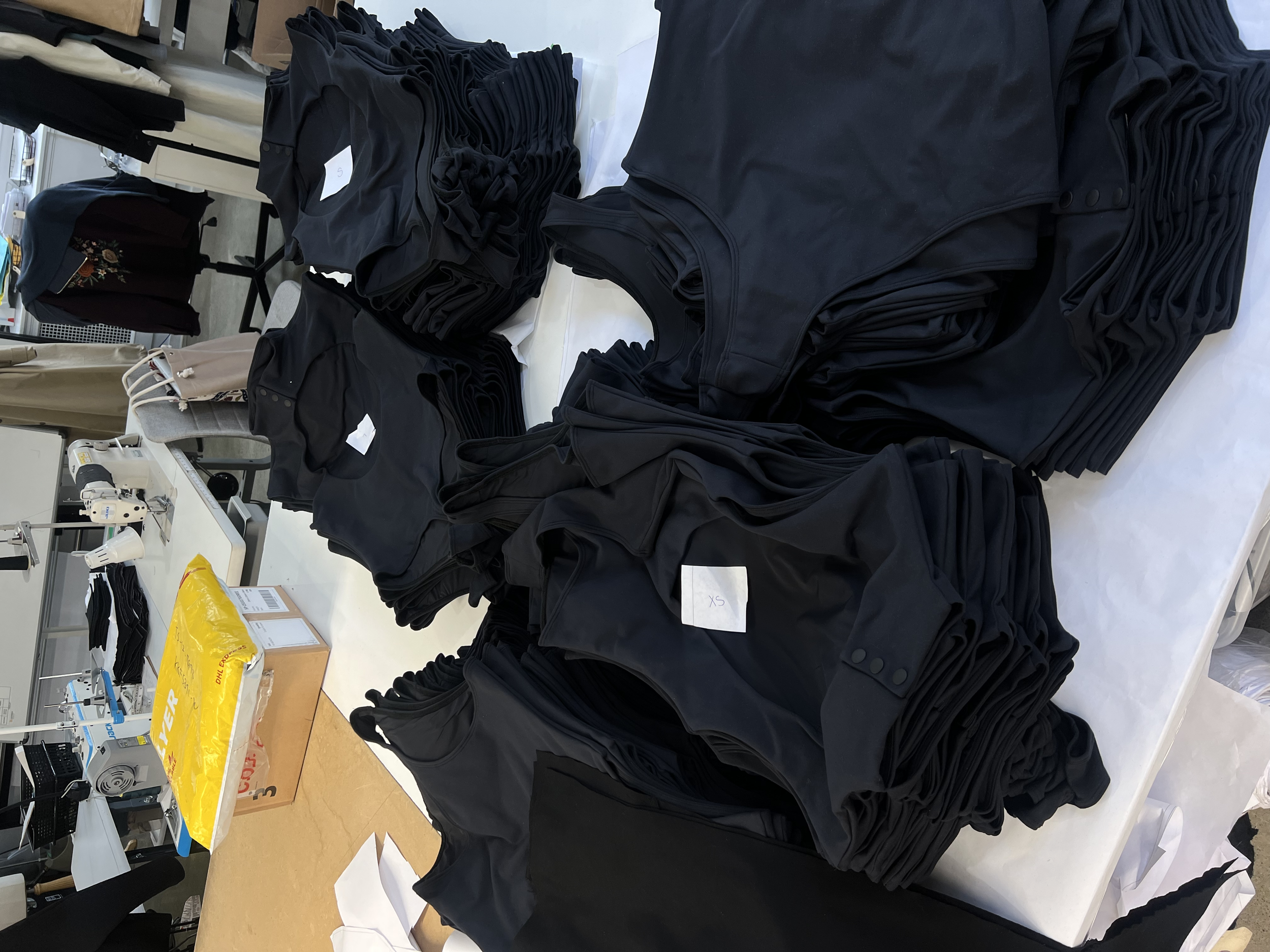Black sleeveless tops or shirts, folded into piles, labeled with size tags 'XS' and '5', on a white table in a manufacturing or packaging setting.