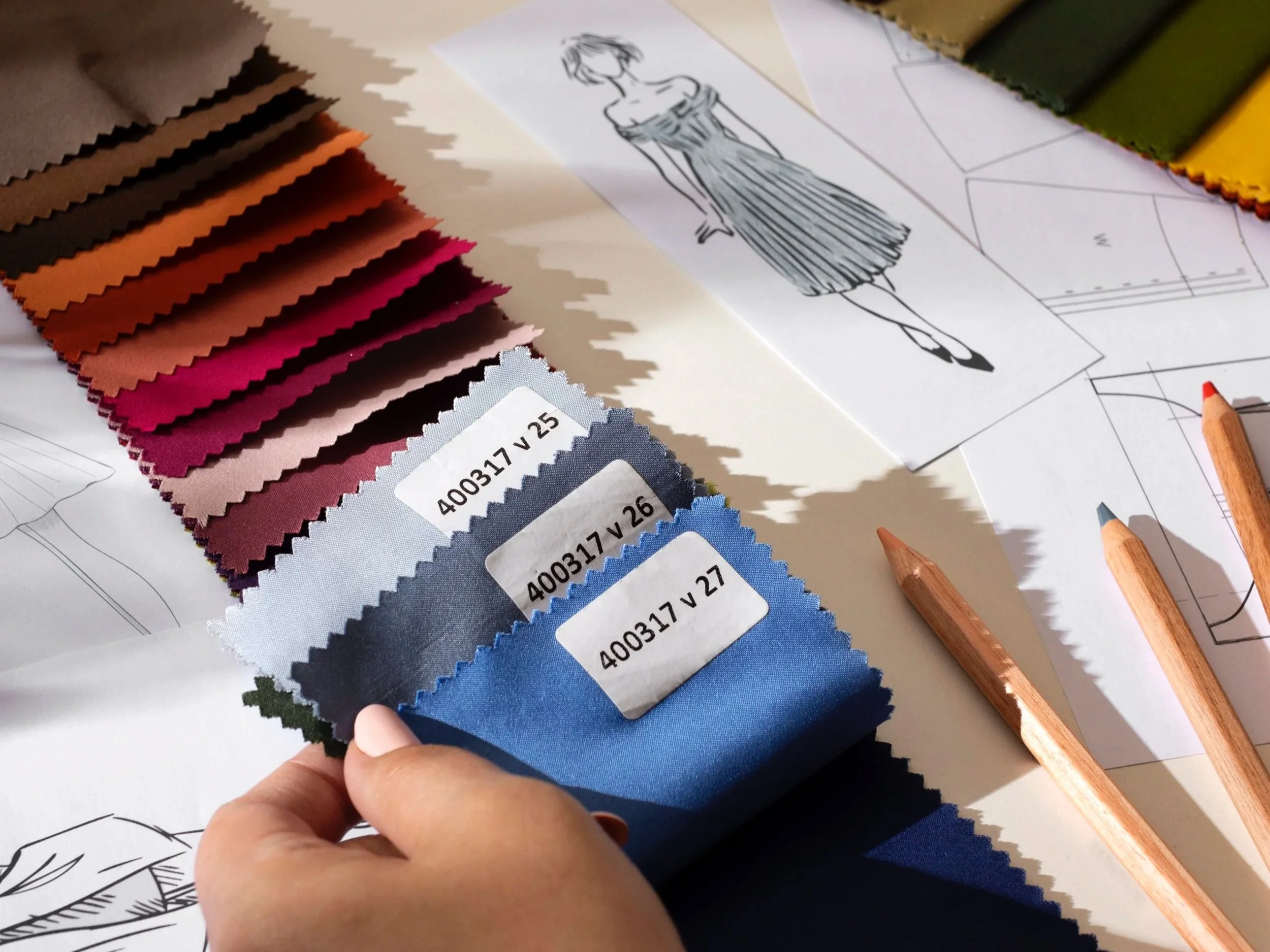 Color swatches for fabric or material samples in shades of pink, blue, green, yellow, and brown, with sketches of a woman in a dress and fabric job labels on the table.