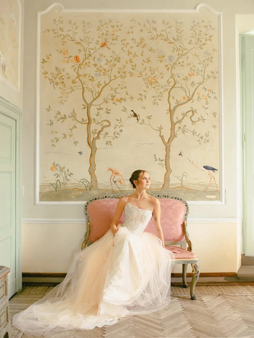 A woman in a wedding dress sitting on a vintage pink and gold sofa in a room with a large decorative wall mural of trees, birds, flowers, and water.