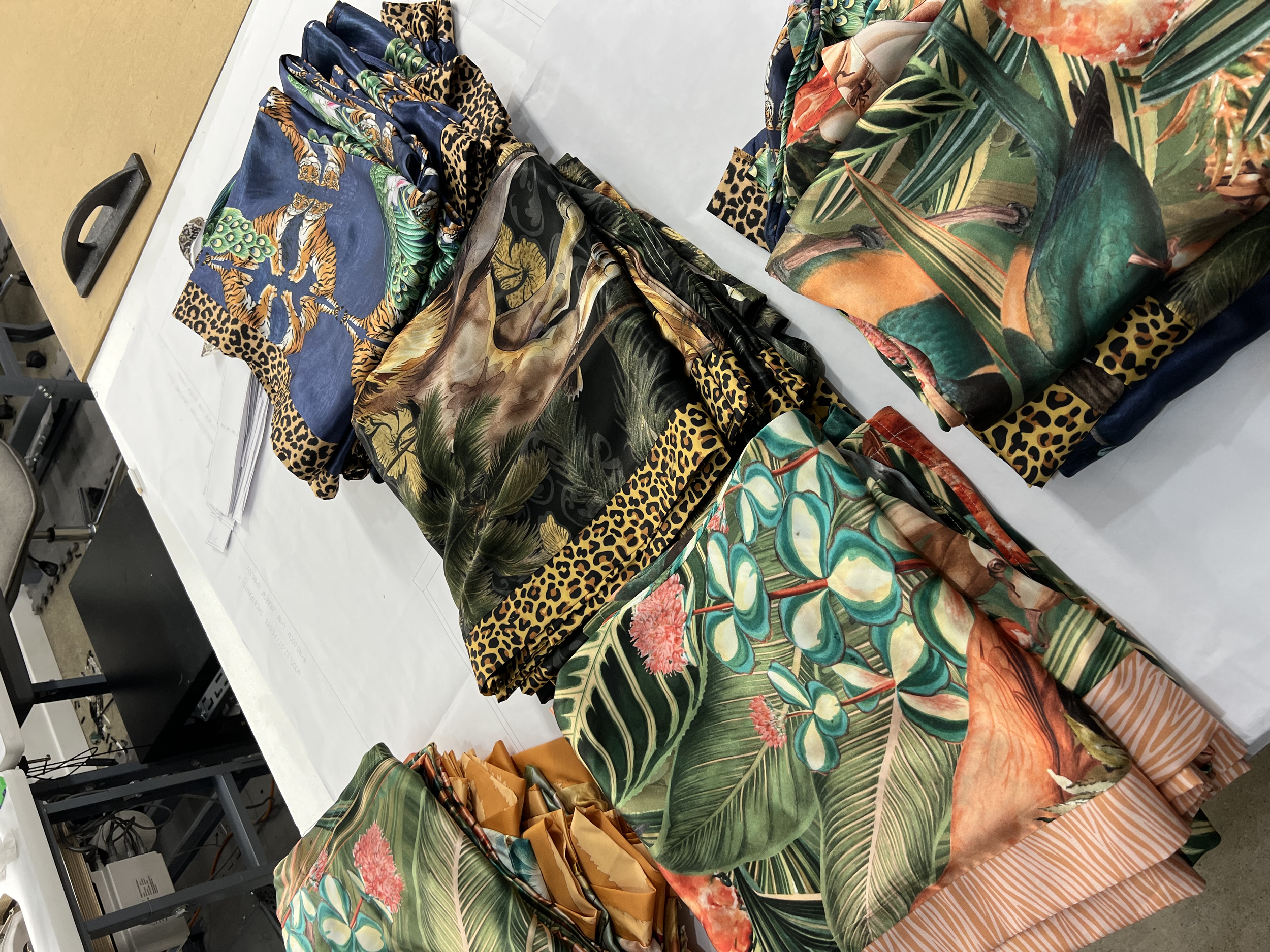 Colorful fabric with jungle and animal prints, including tigers, peacocks, and exotic plants, neatly folded and arranged on a white table.