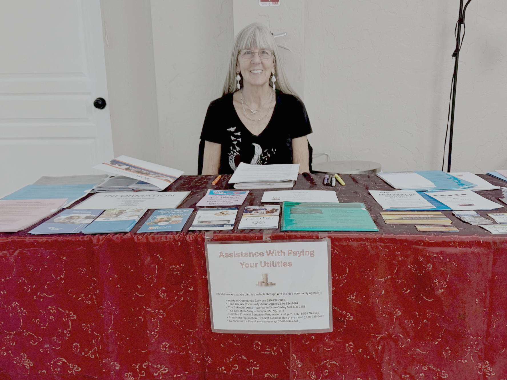 A woman sitting behind a table with various informational pamphlets and flyers about assistance with paying utilities, on a red tablecloth, in an indoor setting.