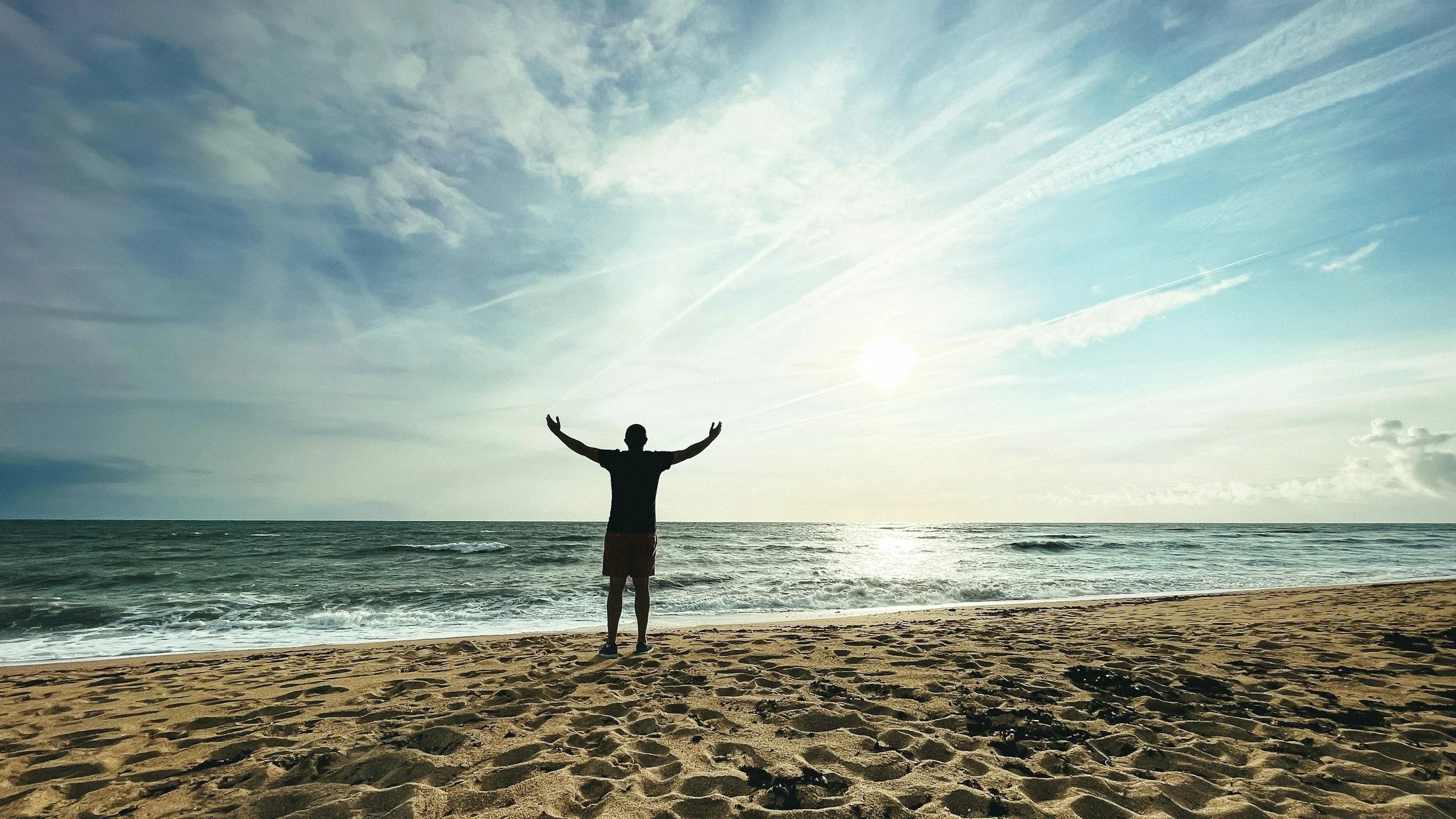 Person standing on a sandy beach with arms raised, facing the ocean during sunset or sunrise, with scattered clouds and a mostly clear sky.