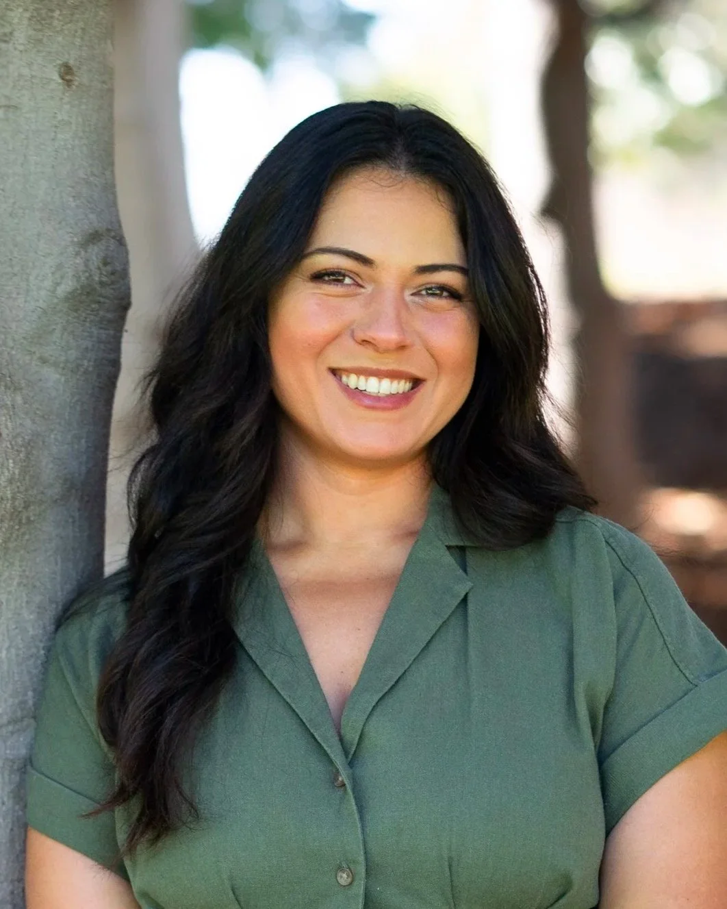 A woman with long black hair, wearing a green shirt, smiling outdoors next to a tree.