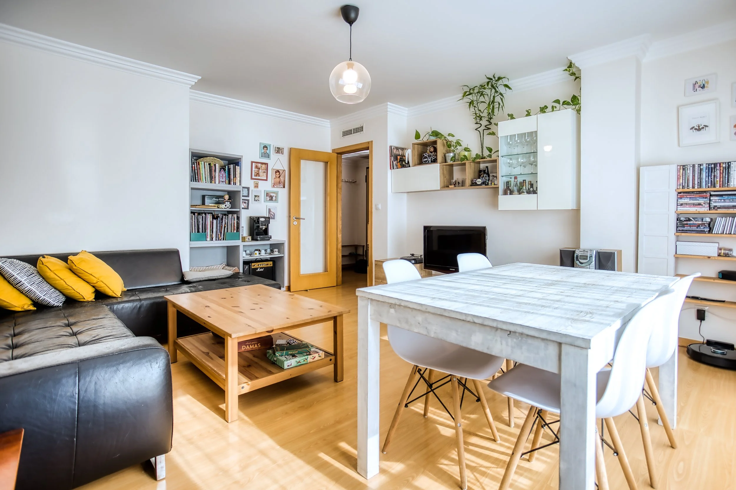 Sala de estar com sofá preto, mesa de centro de madeira, televisão, mesa de jantar com cadeiras brancas, prateleiras com livros, plantas, teto com luminária e chão de madeira.