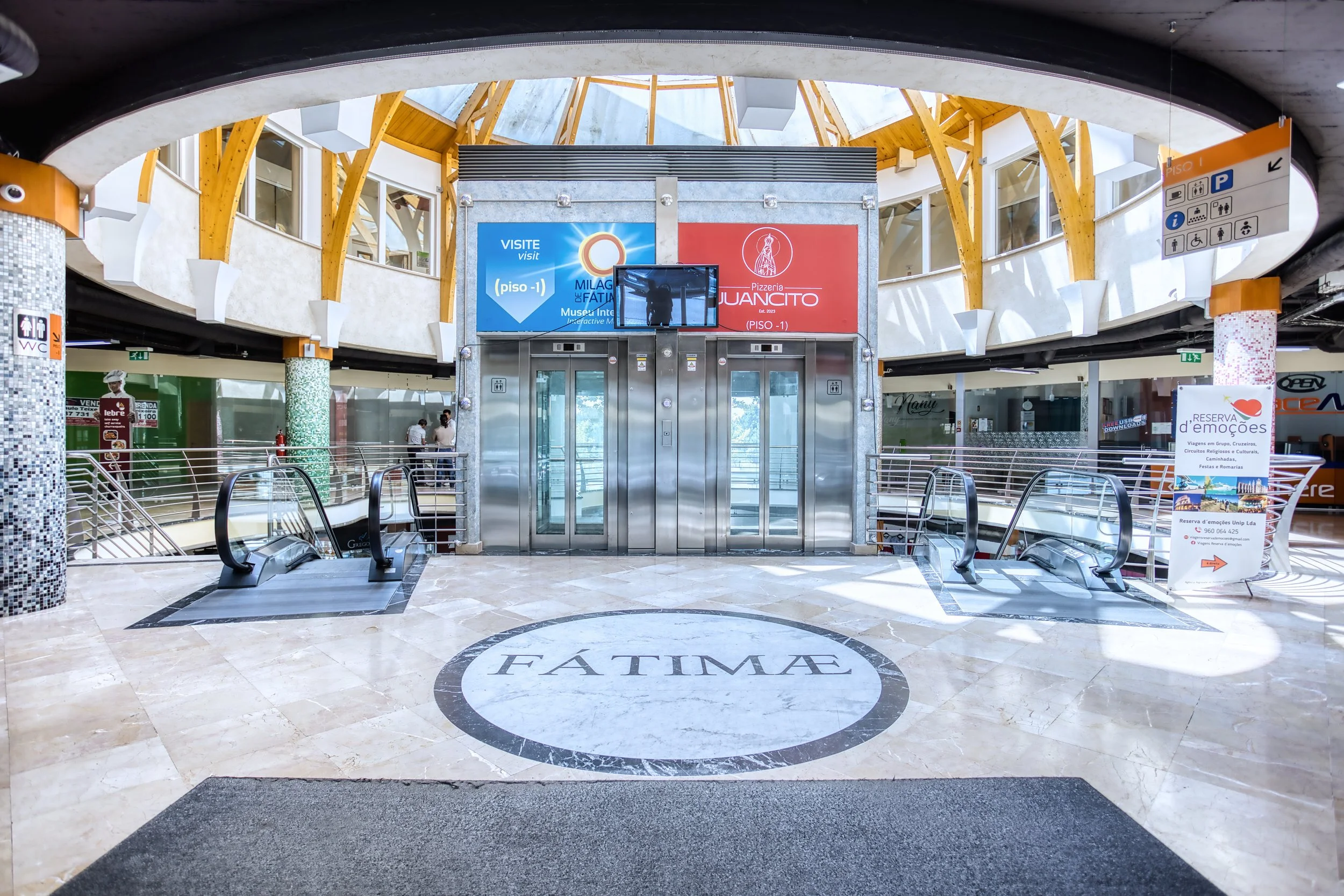 Entrada do shopping FÁTIMA com elevadores, escadas rolantes e decoração moderna com teto de madeira e vidro