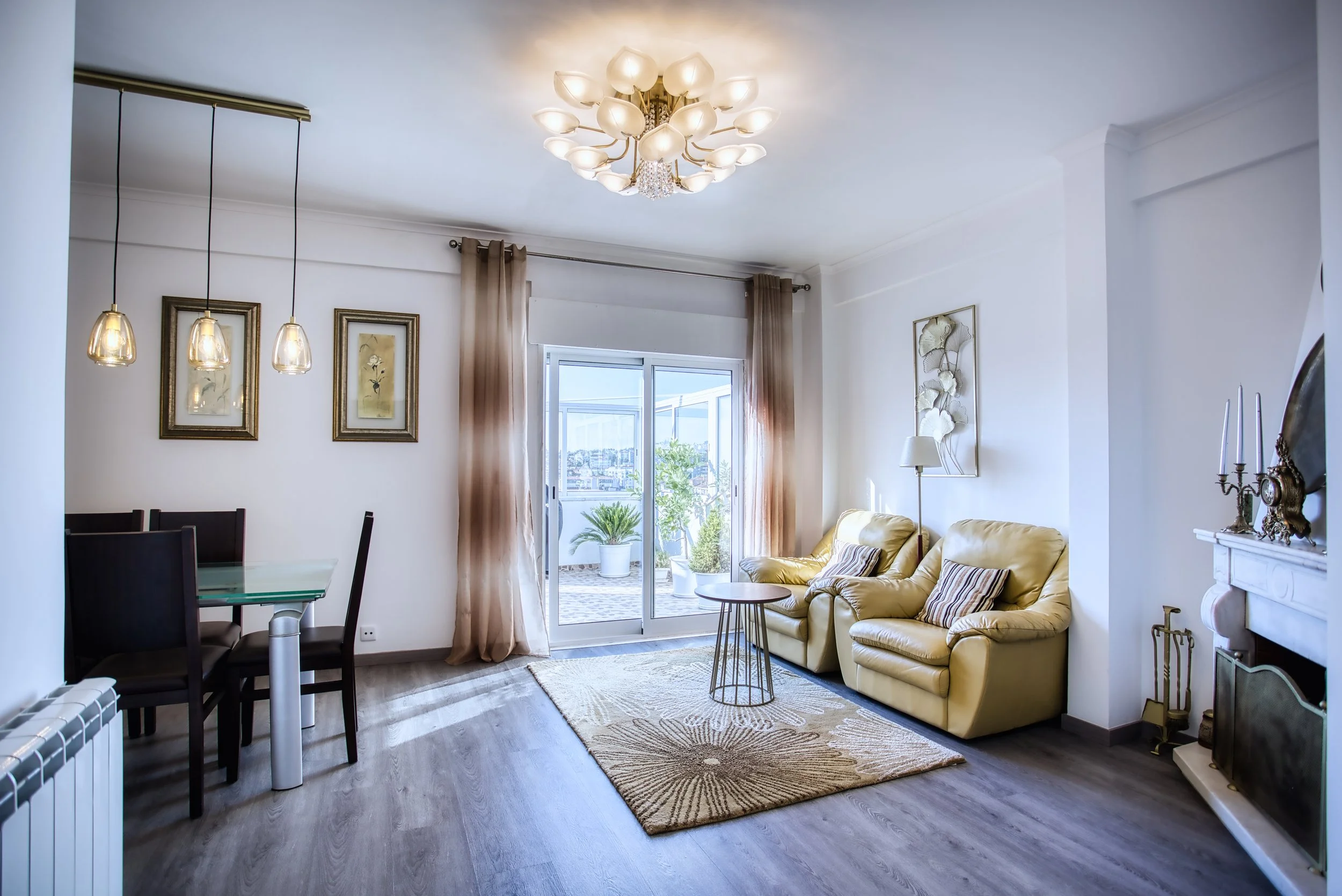 Sala de estar com sofá de couro amarelo, mesa de centro, tapete, quadro na parede, cortinas, iluminação de teto, janelas de vidro com porta de correr levando a um espaço externo com plantas.