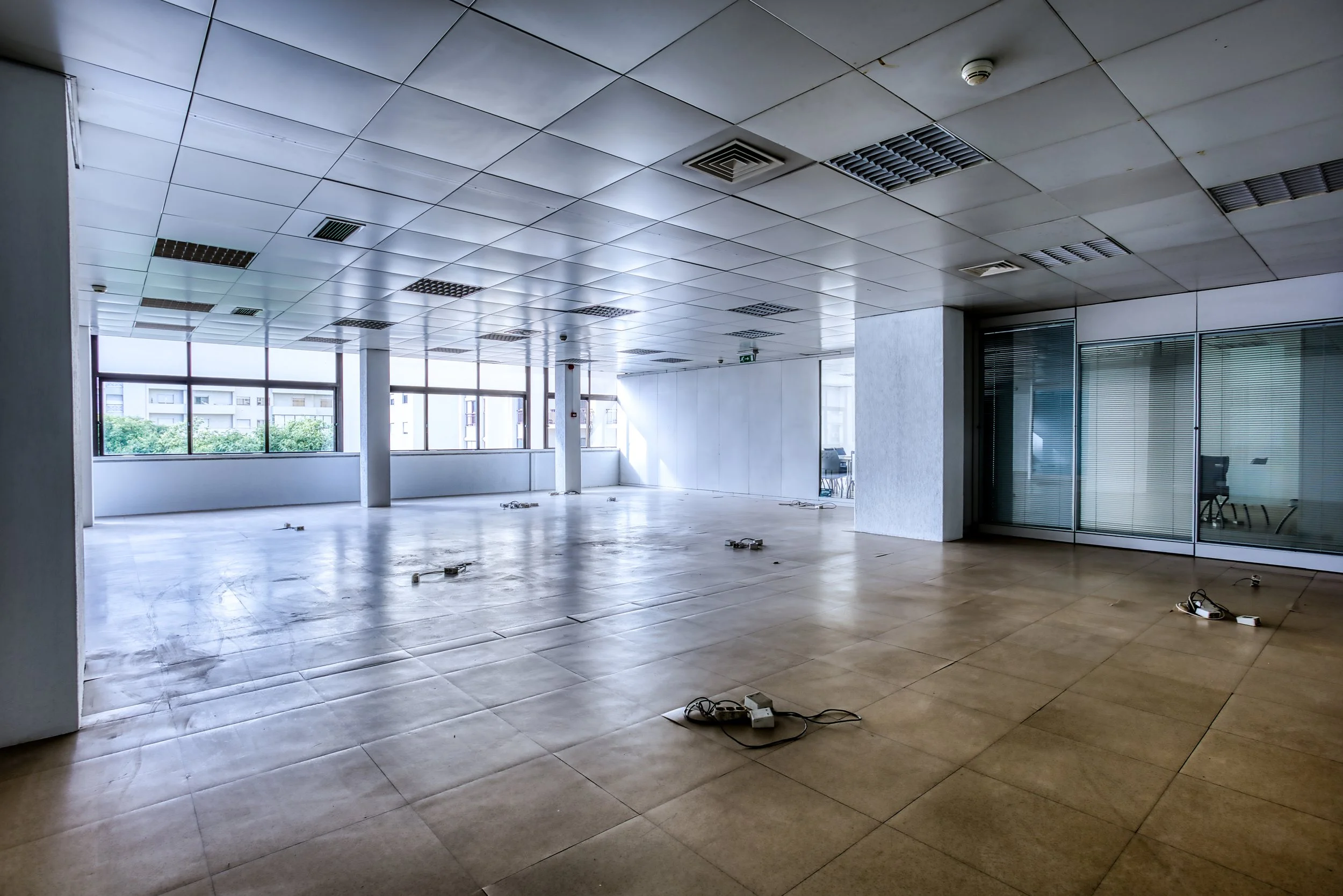 Sala de escritório vazia com piso de azulejos, várias tomadas no chão, janelas grandes e teto com luminárias de iluminação, parcialmente iluminada.