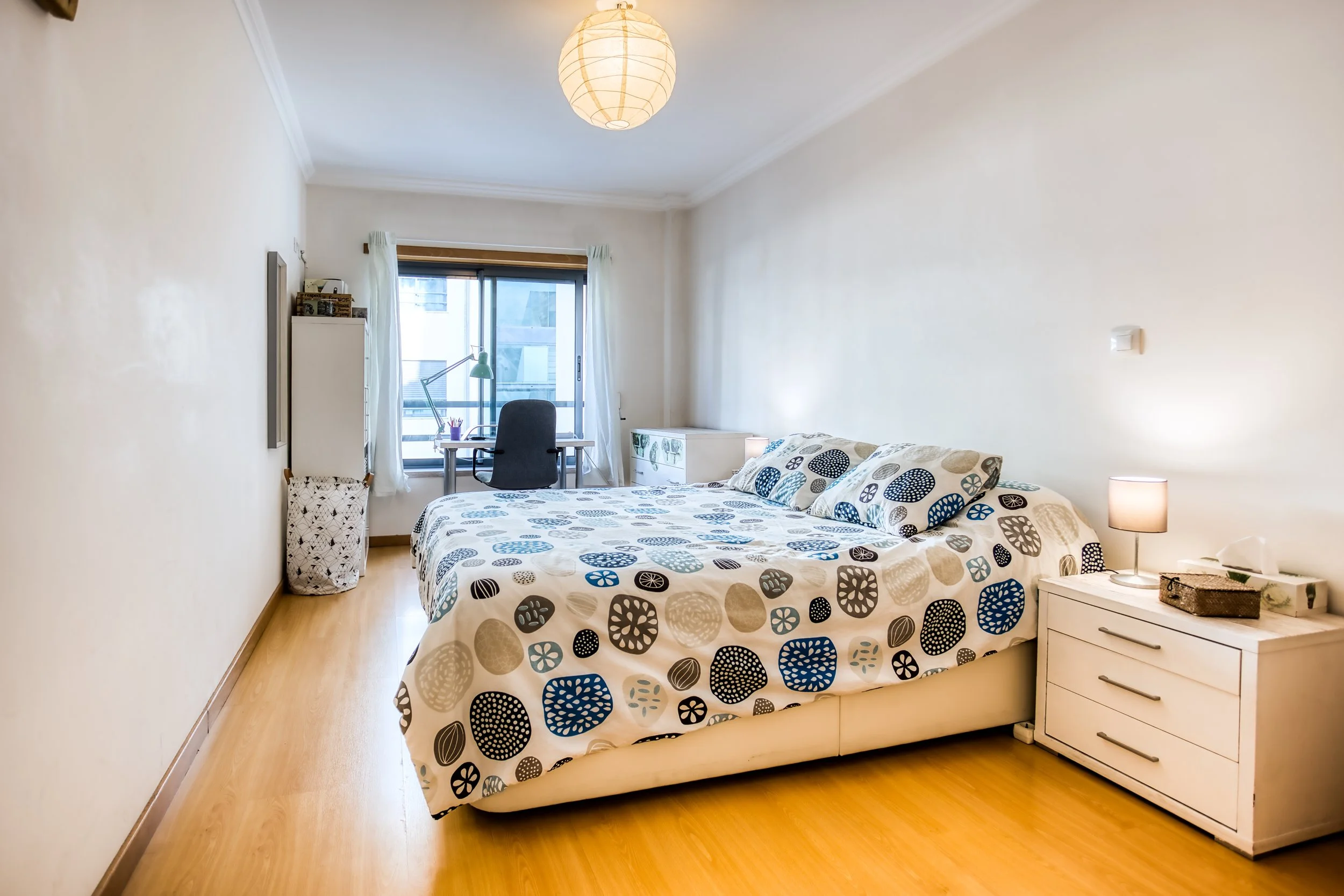 Quarto com cama de casal com roupa de cama com padrão de círculos em cores bege, azul e preto, ao lado de uma penteadeira branca com uma luminária, ao fundo uma janela grande com cortinas brancas, uma cadeira de escritório e uma mesa de estudo com lu