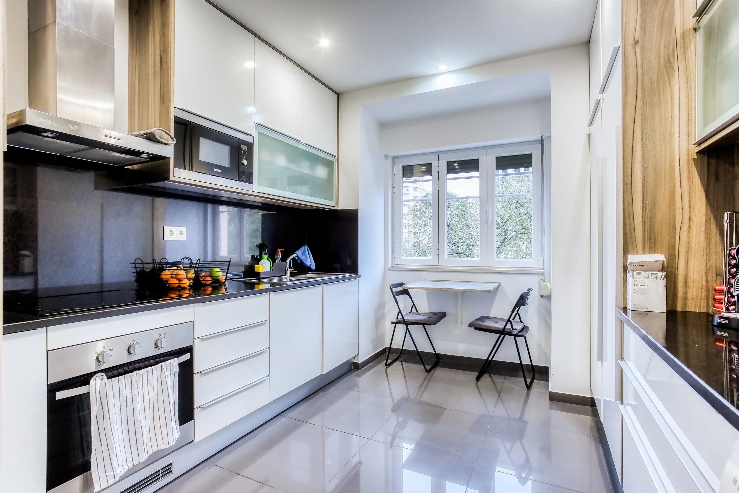 Cozinha moderna com armários brancos, pia, bancada preta, janela grande, duas cadeiras pequenas na janela, eletrodomésticos, e uma parede de madeira com itens.