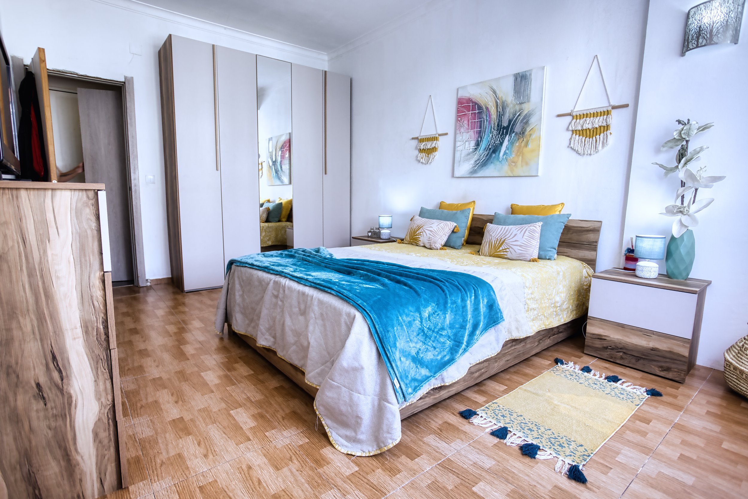 Quarto com cama de madeira, roupas de cama amarelas e azuis, paredes brancas decoradas com quadros e têxteis pendurados. Cómoda de madeira no lado esquerdo, armário de portas brancas com espelho na parede ao fundo, floreira com plantas no lado direit