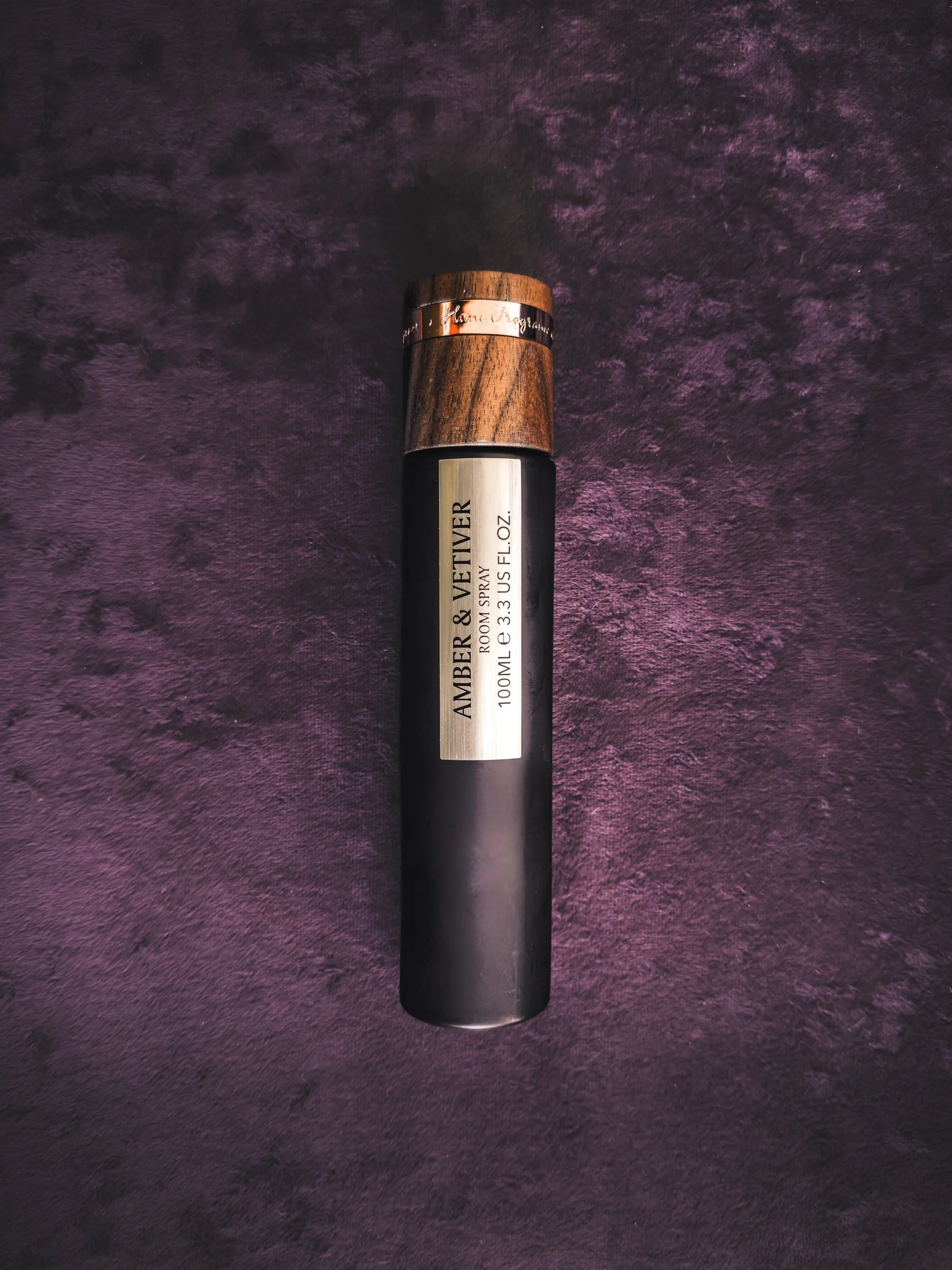 Frasco de perfume com tampa de madeira escura e rótulo prata escuro, sobre fundo roxo