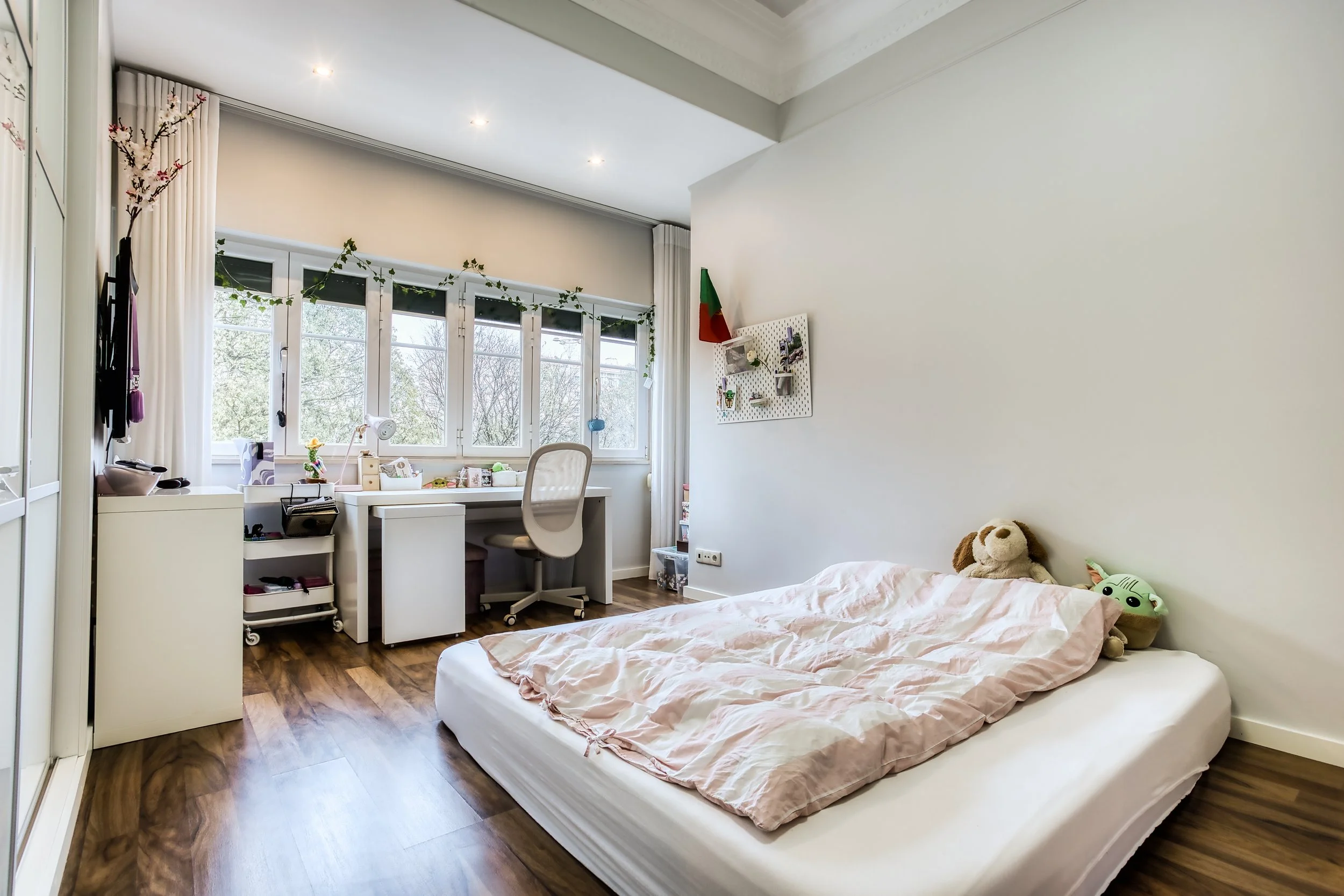 Quarto com cama no chão, roupas de cama rosadas, dois pelúcias na cama, uma parede branca com prateleira de parede, mesa de estudo junto às janelas com cadeira, cortinas brancas, piso de madeira, muitas janelas com vista para árvores.