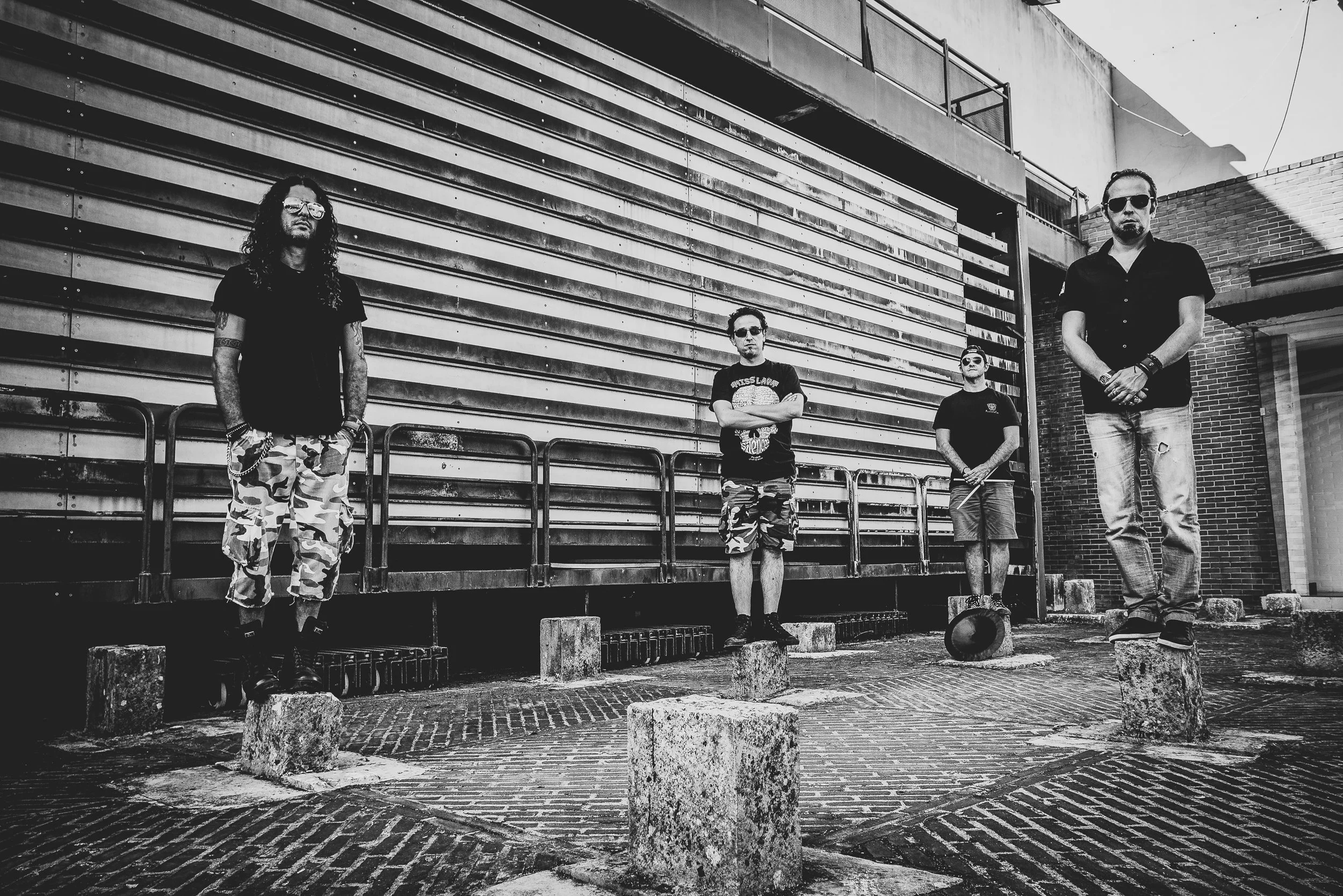 Quatro pessoas vestidas casualmente posando em uma rua urbana ao fundo de uma parede de painéis de metal.