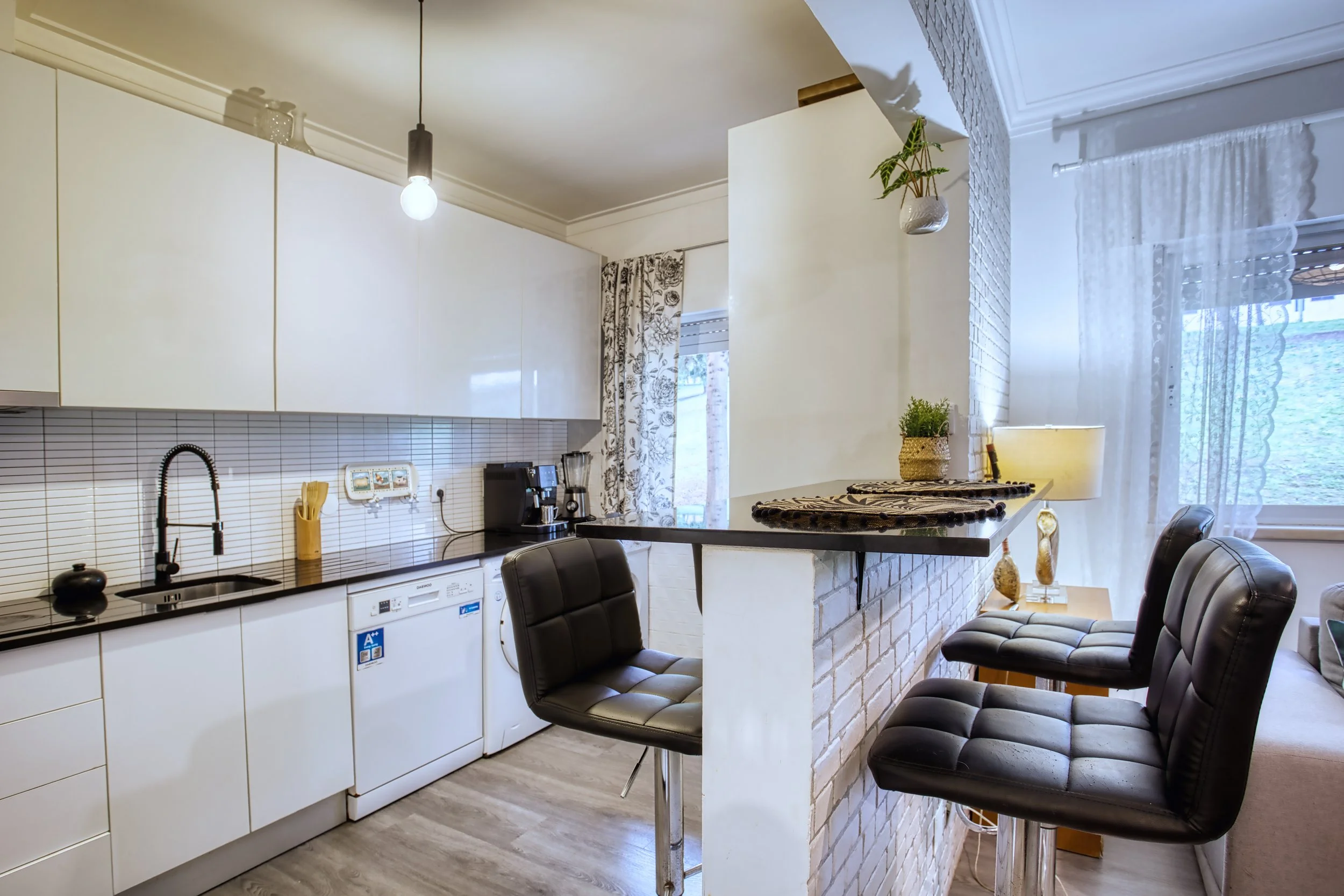 Cozinha moderna com varanda, pia preta, armários brancos, eletrodomésticos, balcão com duas cadeiras pretas, plantas decorativas, cortinas e iluminação