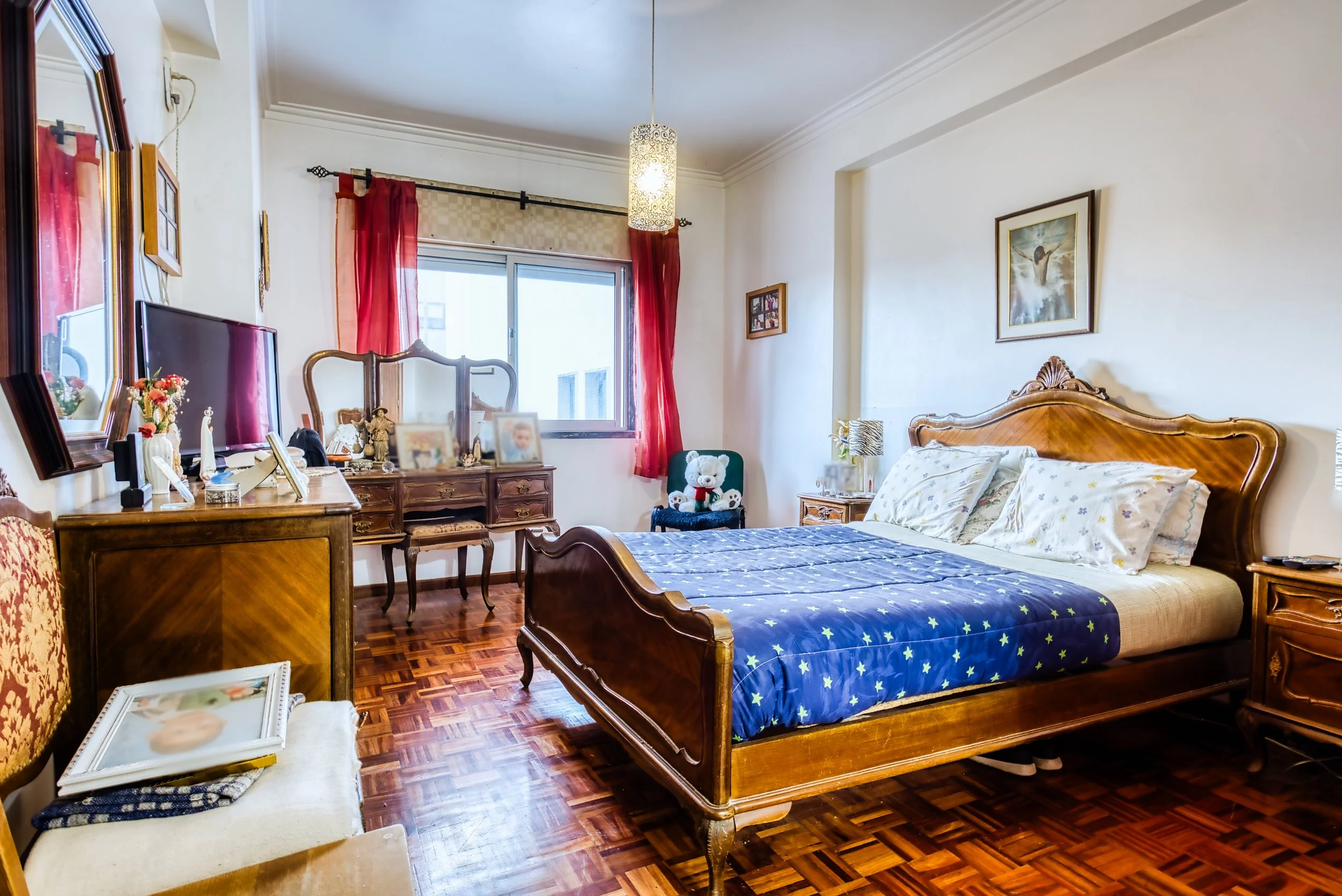 Quarto com cama de madeira, roupas de cama de estrelas, paredes brancas, janela com cortinas vermelhas, móveis antigos, televisão, decoração com fotos e objetos pessoais, piso de parquet, abajur, quadro na parede.