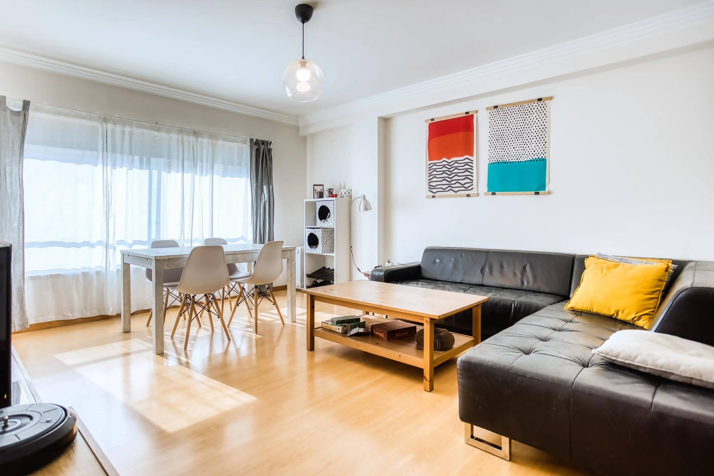 Sala de estar com sofá preto, mesa de centro de madeira, tapete cinza, poltrona branca, mesa de jantar branca com cadeiras brancas, parede branca com duas obras de arte coloridas, cortinas brancas e cinza, piso de madeira, luminária pendente de vidro