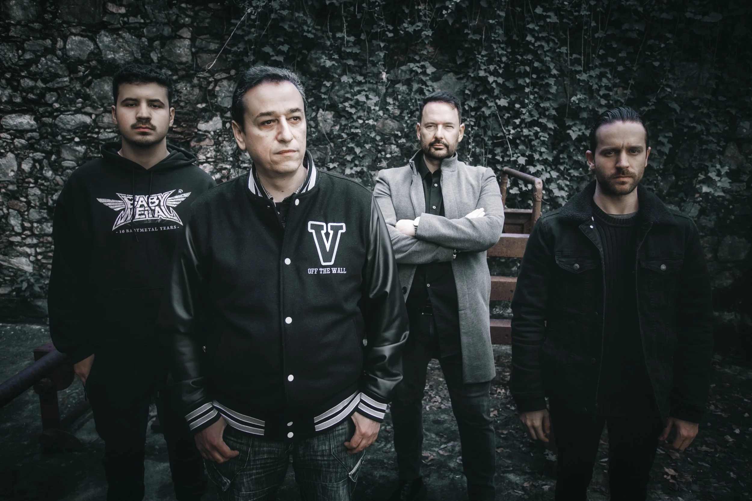 Quatro homens em uma parede de pedra com plantas ao fundo, alguns com braços cruzados, todos com expressão séria e roupas escuras.