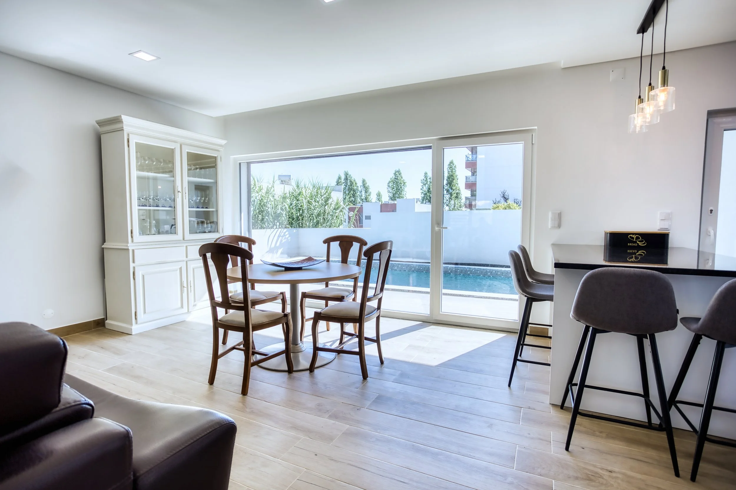 Sala de jantar com mesa redonda de madeira e quatro cadeiras, ao fundo uma janela grande voltada para uma piscina e jardim externo, parede branca, com um lustre de lâmpadas pendurado.